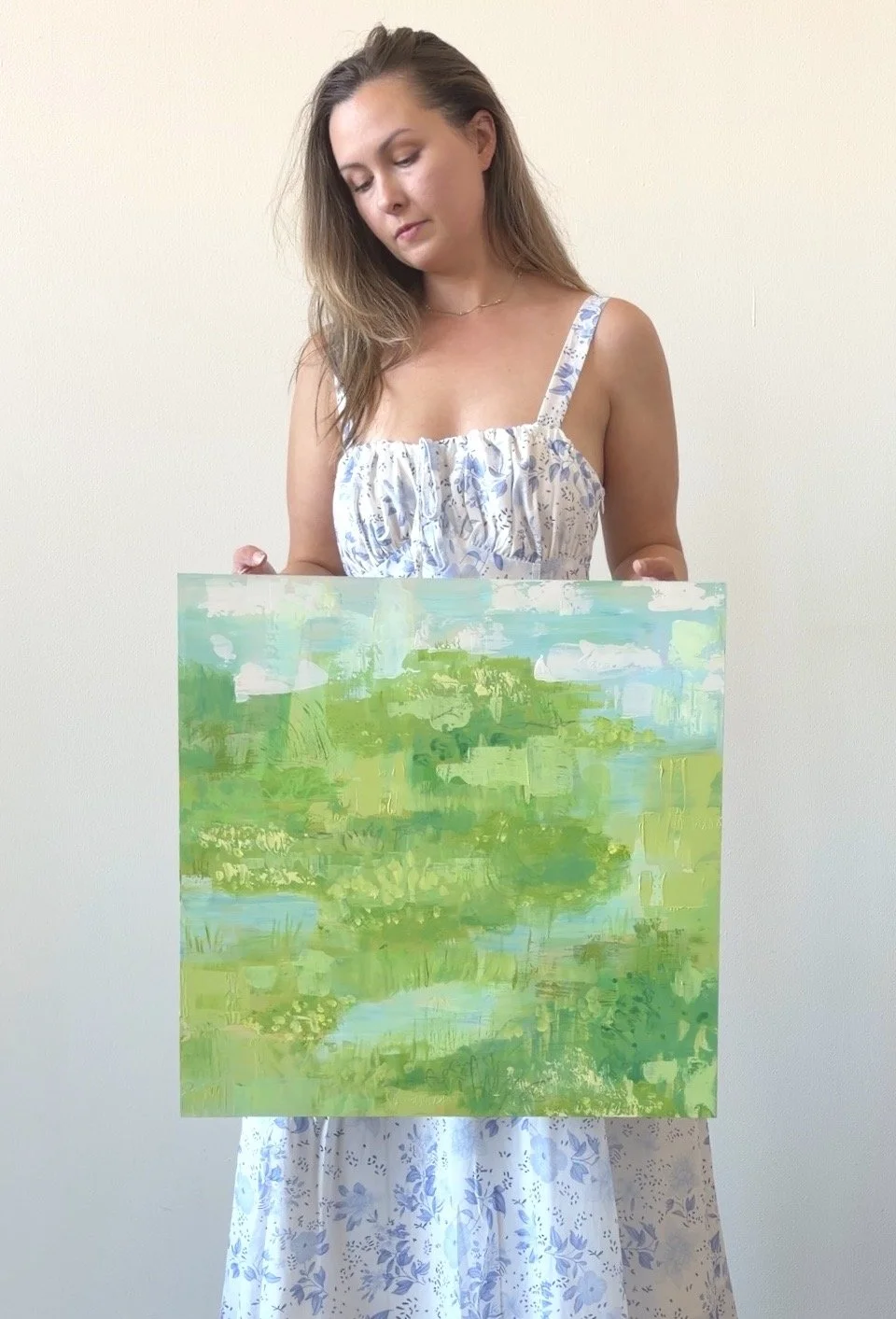A picture of Kellie, the artist, in a blue and white floral dress holding an abstract landscape painting. The painting is a mix of light blues, greens, and white and evokes a sense of calm.