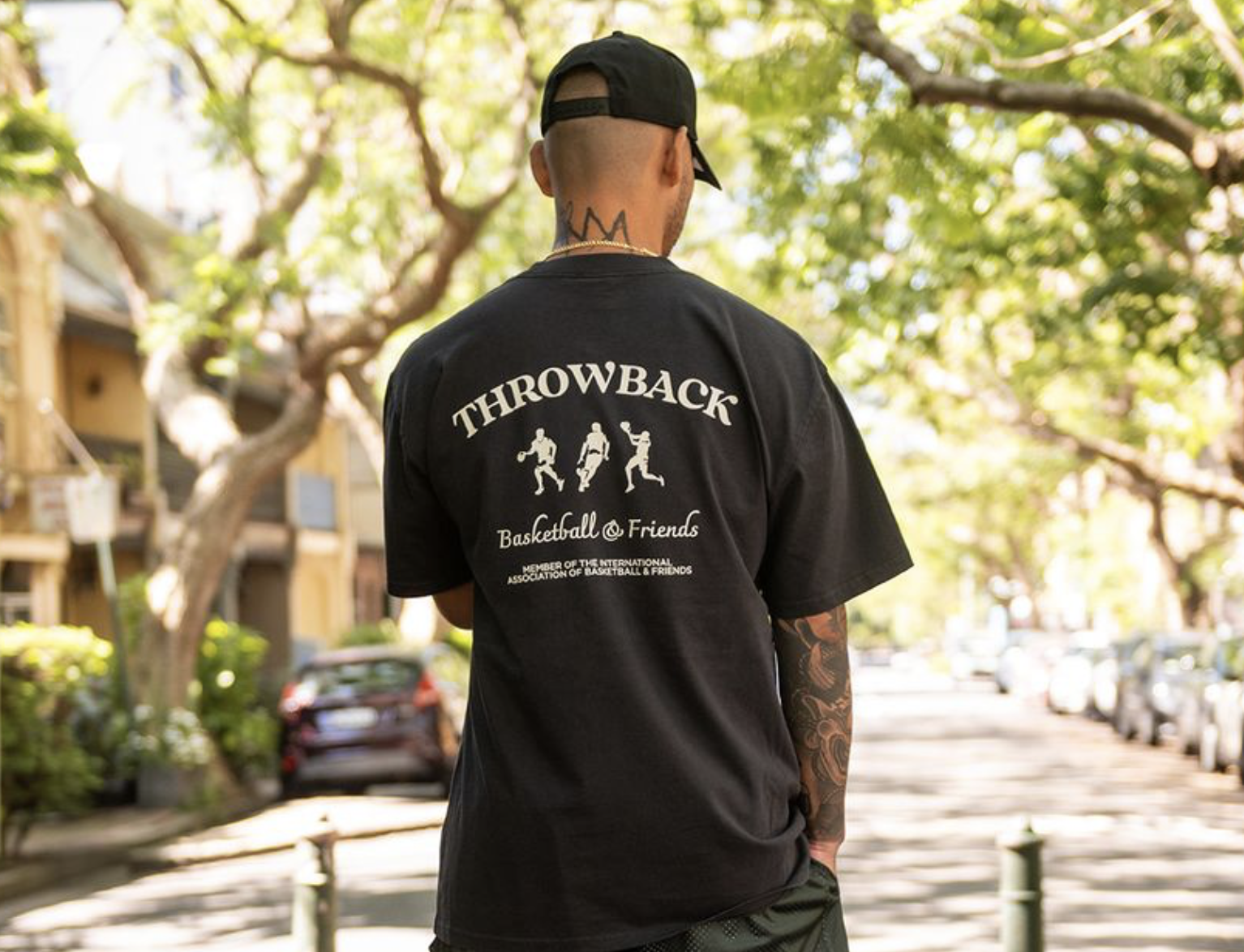 Man with a tattooed arm wearing a black cap and a black T-shirt with "THROWBACK Basketball @ Friends" graphic standing outdoors on a tree-lined street.