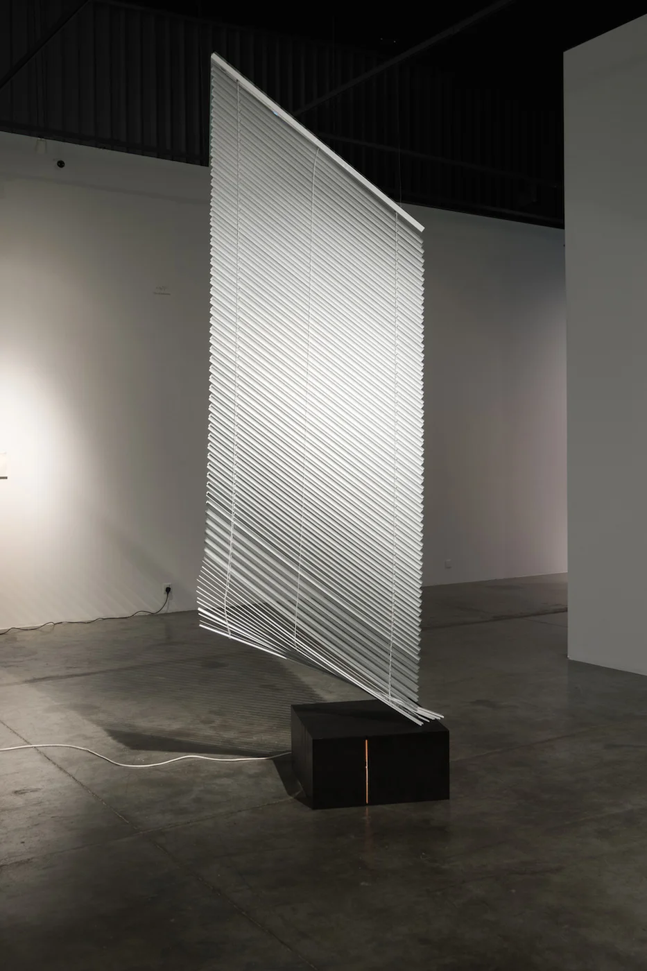 Vittorio Santoro Good-Bye Darkness, V 2012 Venetian blind (250 x 120 cm), suspension devices, steel wires, black steel base (30 x 60 x 60 cm), light bulb with base, electric cord with plug Dimensions variable The suspended venetian blind rests partly on a rectangular black steel base. The base comprises vertical slits on two of its sides through which rays of light escape. They stem from an electric light bulb invisibly placed in the interior. The measurements of the base echoes Concrete Tape Recorder Piece (1968) by Bruce Nauman.