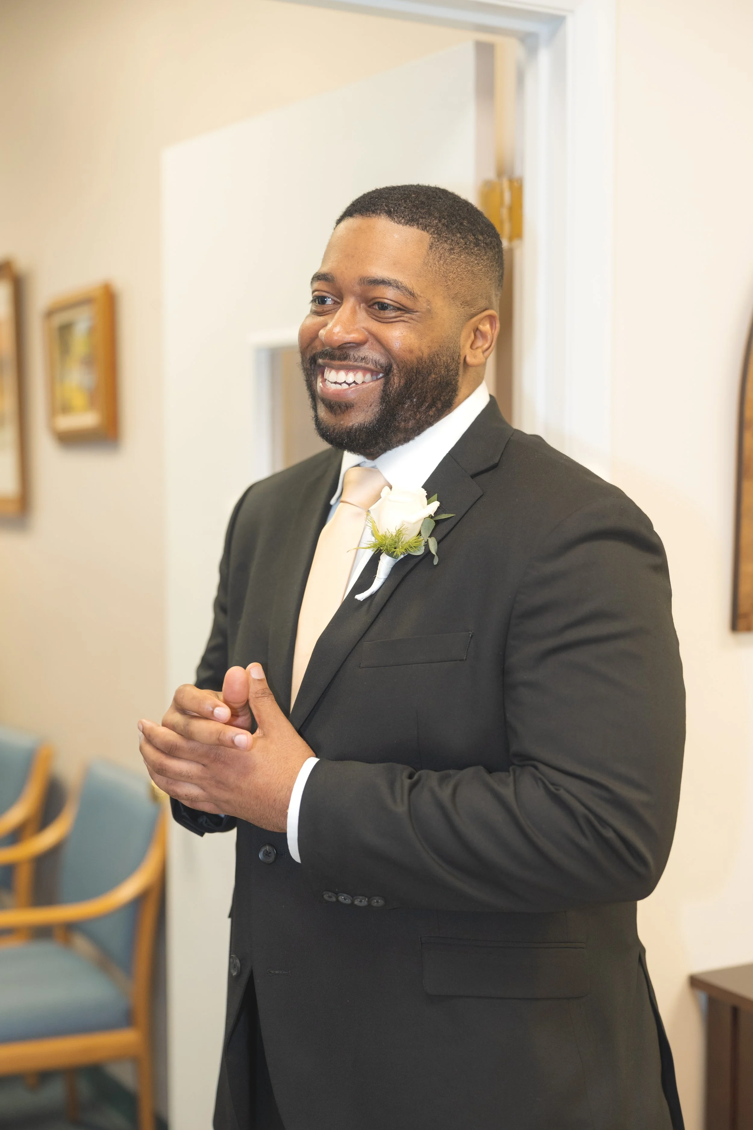 A smiling man in a black suit with a white shirt and beige tie, wearing a white rose boutonniere, standing indoors.