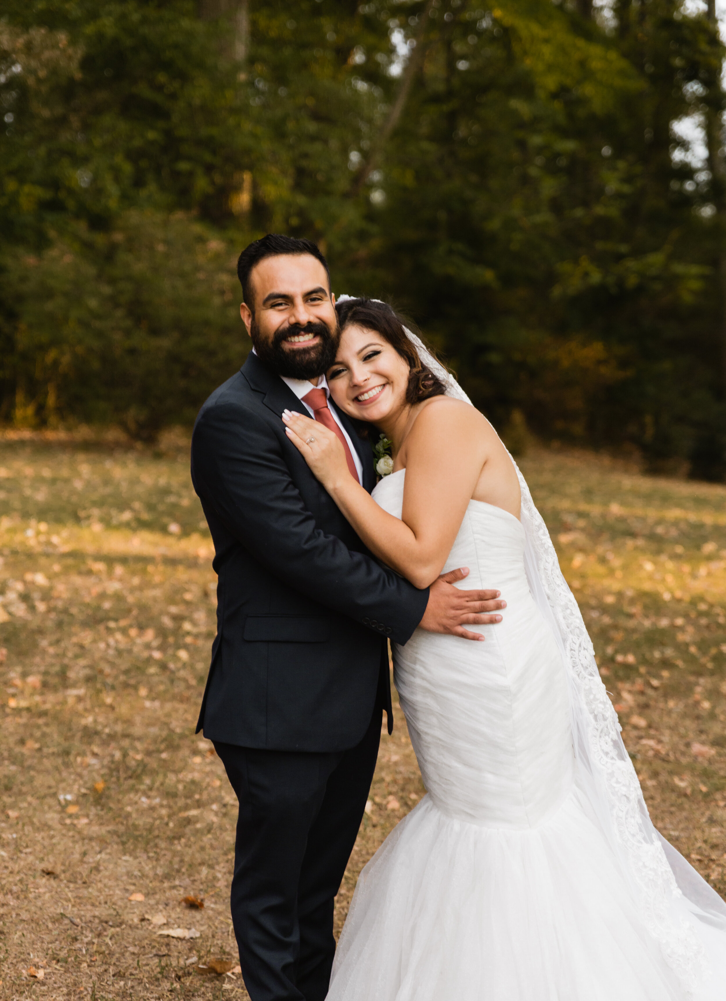 A bride and groom smiling and hugging outdoors in a wooded setting, dressed in wedding attire.