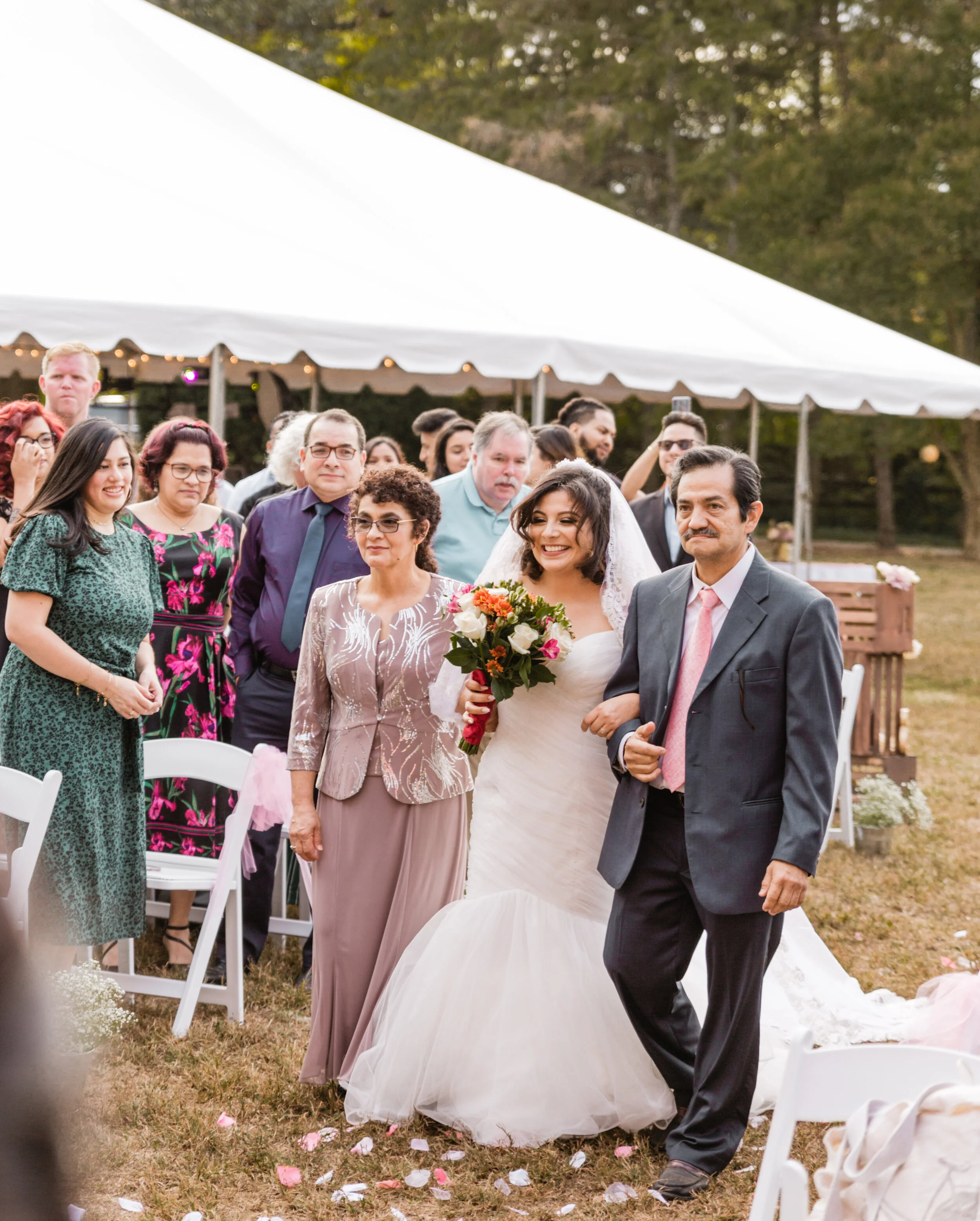 Bride walking down the aisle with her parents, smiling, holding a bouquet of flowers, surrounded by guests at an outdoor wedding reception under a white tent.