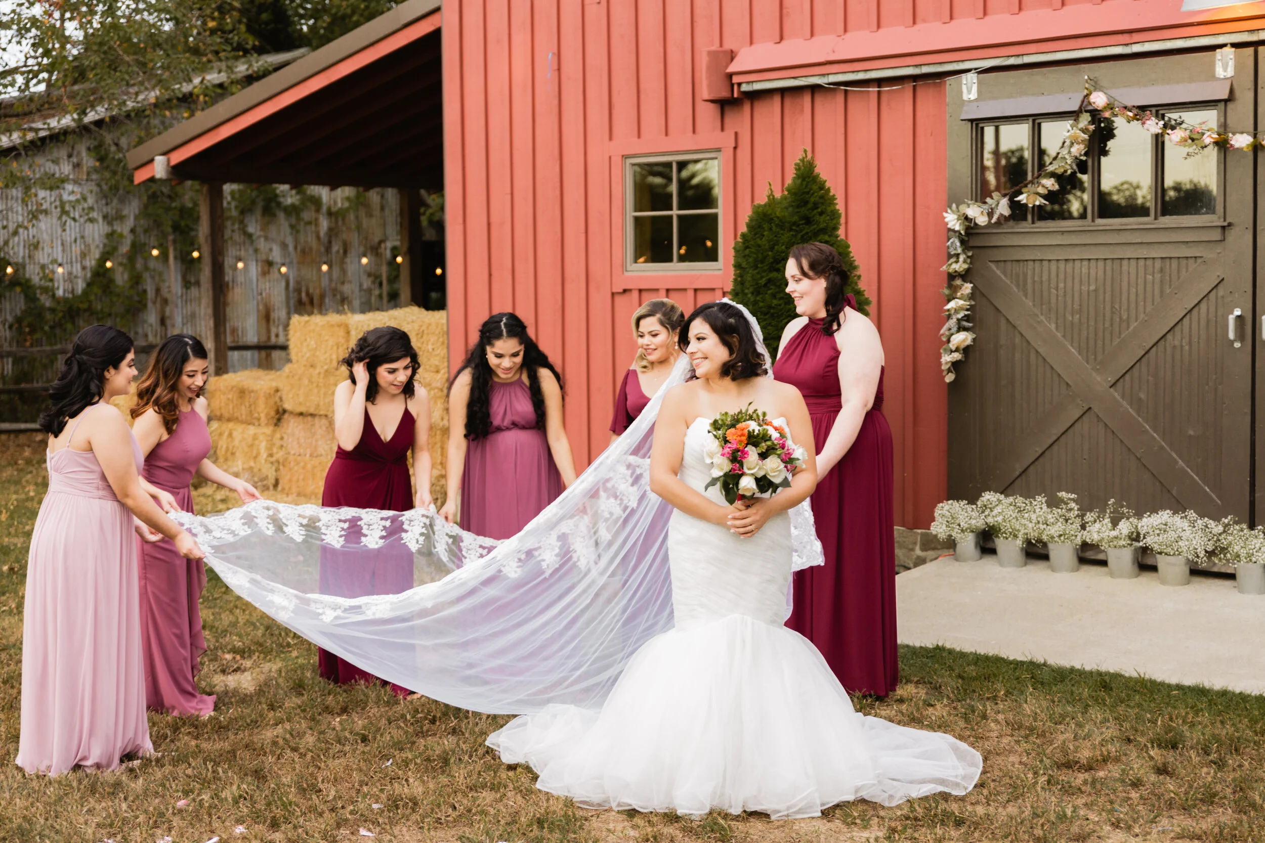 Bride in white wedding gown holding a bouquet, standing on grass, with six bridesmaids in pink and purple dresses holding the train of her veil in front of a red barn decorated with flowers.