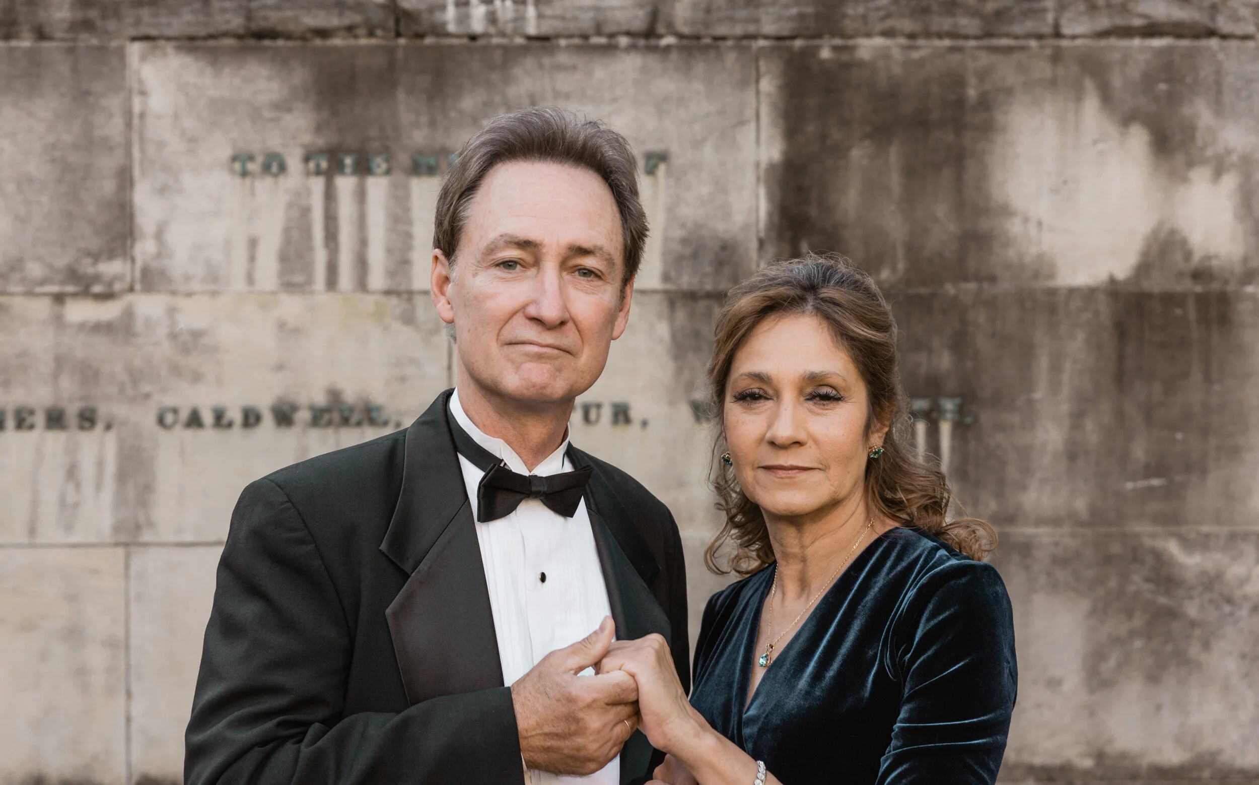 A married couple dressed formally, the man in a tuxedo and the woman in a velvet dress, holding hands in front of a stone monument.