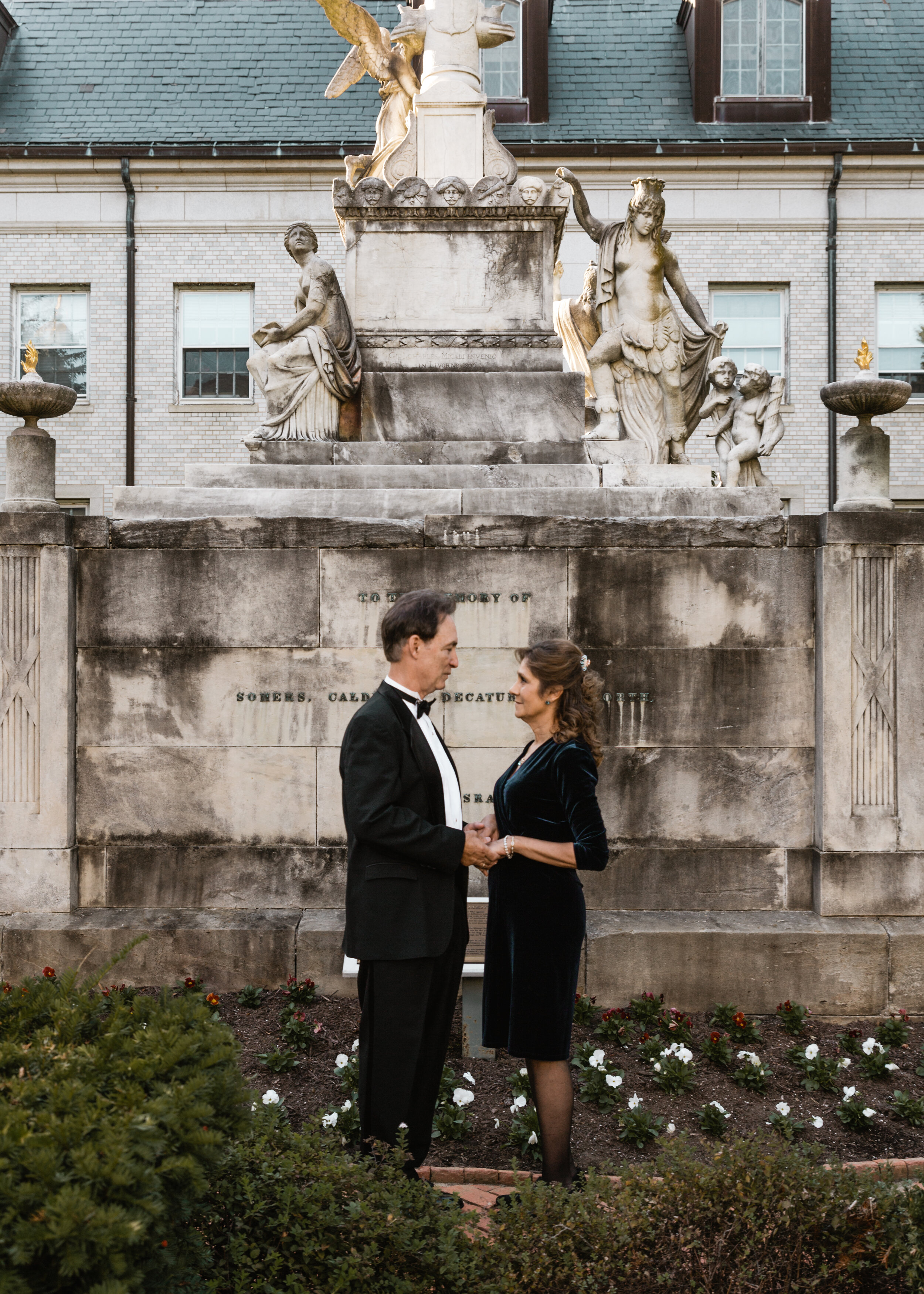 A man and woman dressed in formal attire holding hands and facing each other in front of a historic monument with sculptures, flowers, and a building in the background.