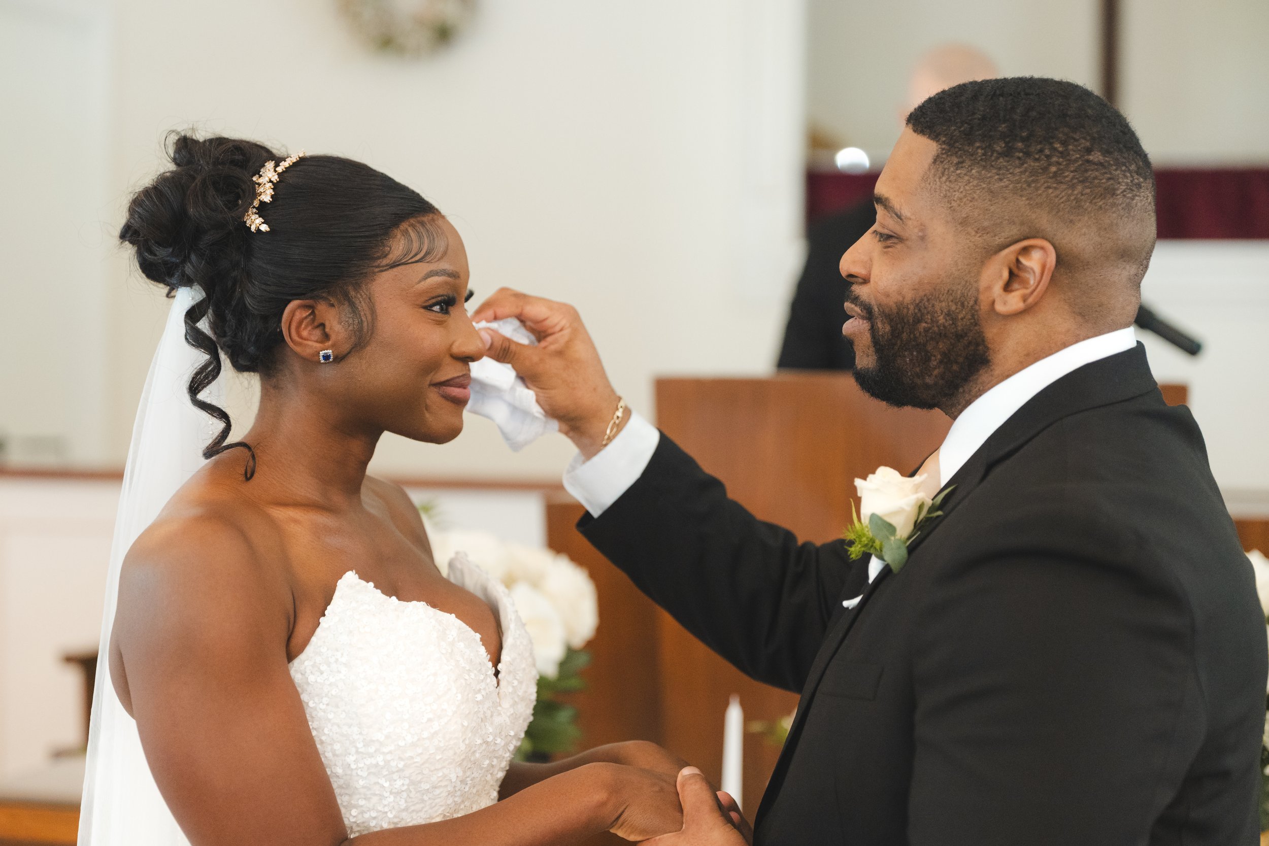 A bride and groom exchanging vows in a wedding ceremony, with the groom gently wiping a tear from the bride's eye.