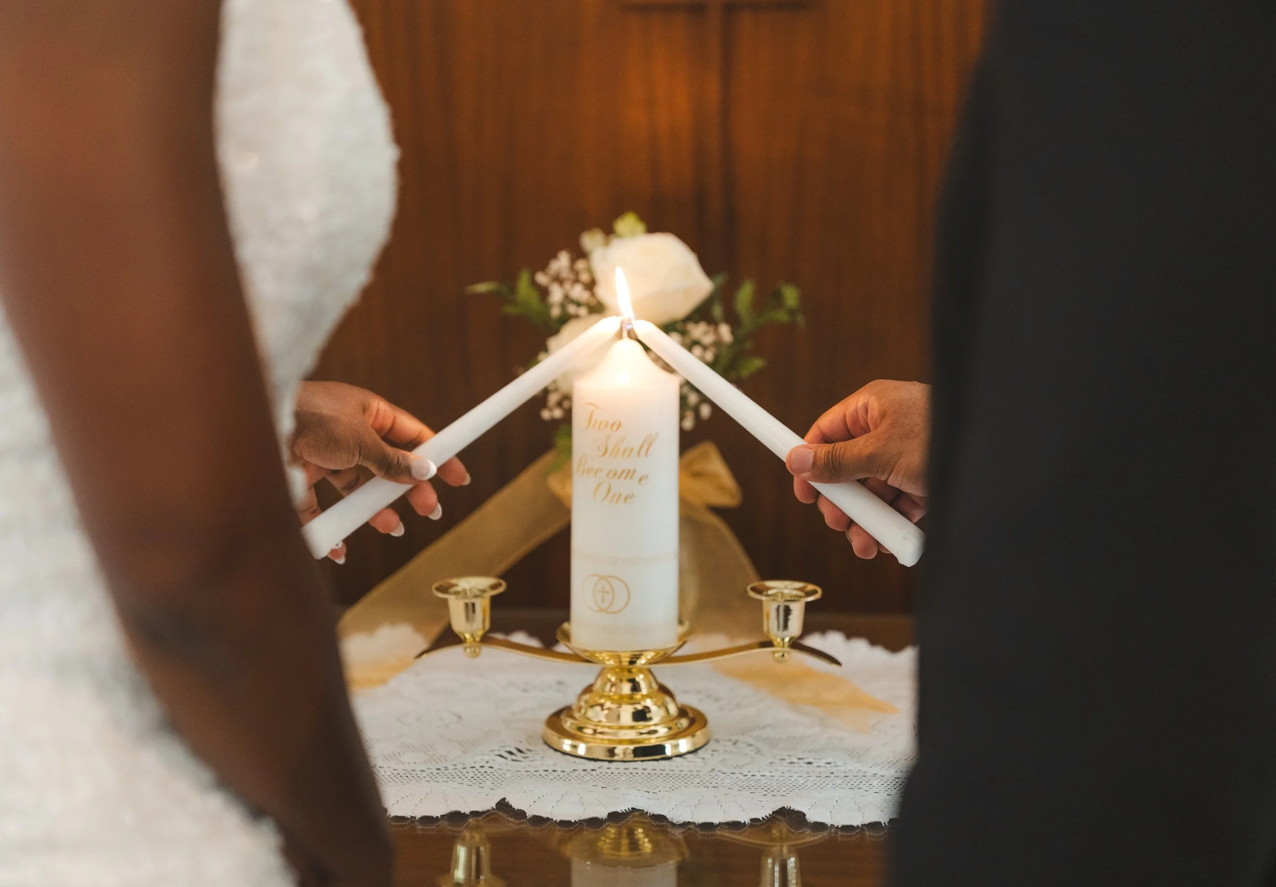 A couple lighting a unity candle during a wedding ceremony with a white candle inscribed "Two Shall Become One" on a gold holder, with floral decorations in the background.