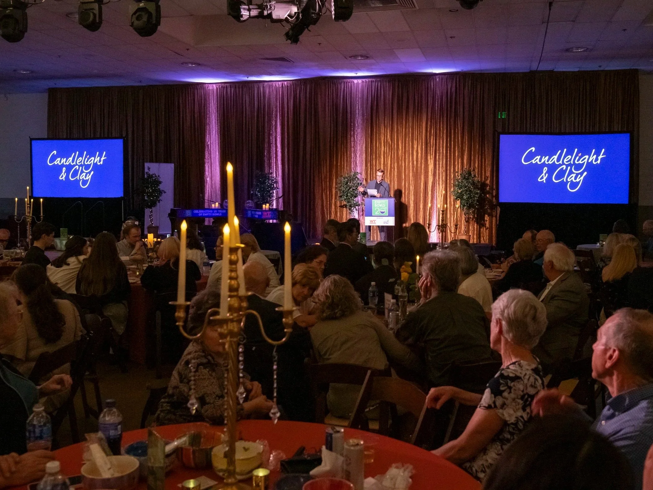 A group of people sitting and listening to a speaker. The speaker stands at a podium and is flanked by two large screens reading "Candlelight & Clay". In the foreground is a gold candelabra.