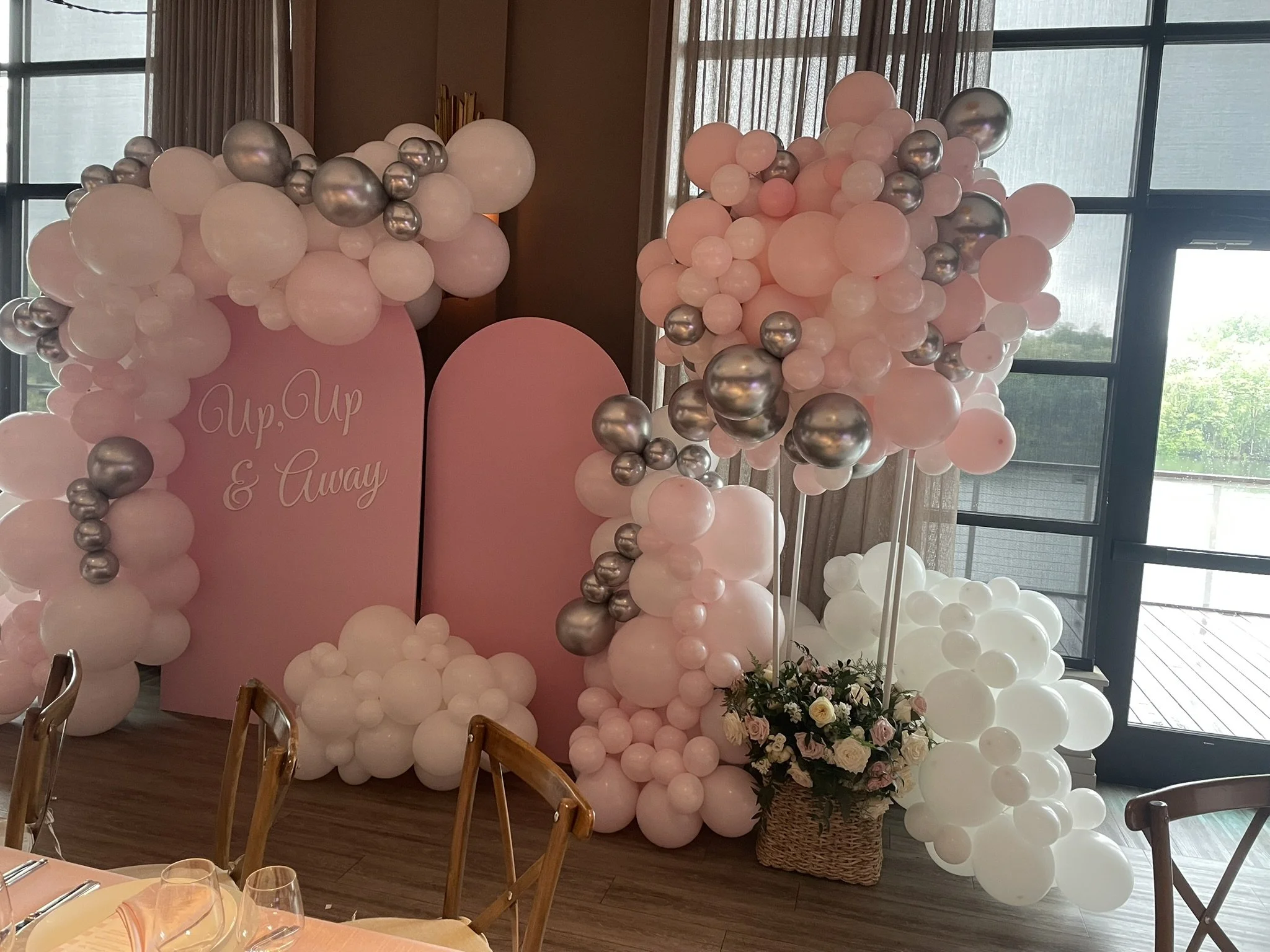 Decorative balloon arrangement with pink and silver balloons, a pink backdrop with cursive writing, and a flower arrangement in a basket, set up indoors near large windows.