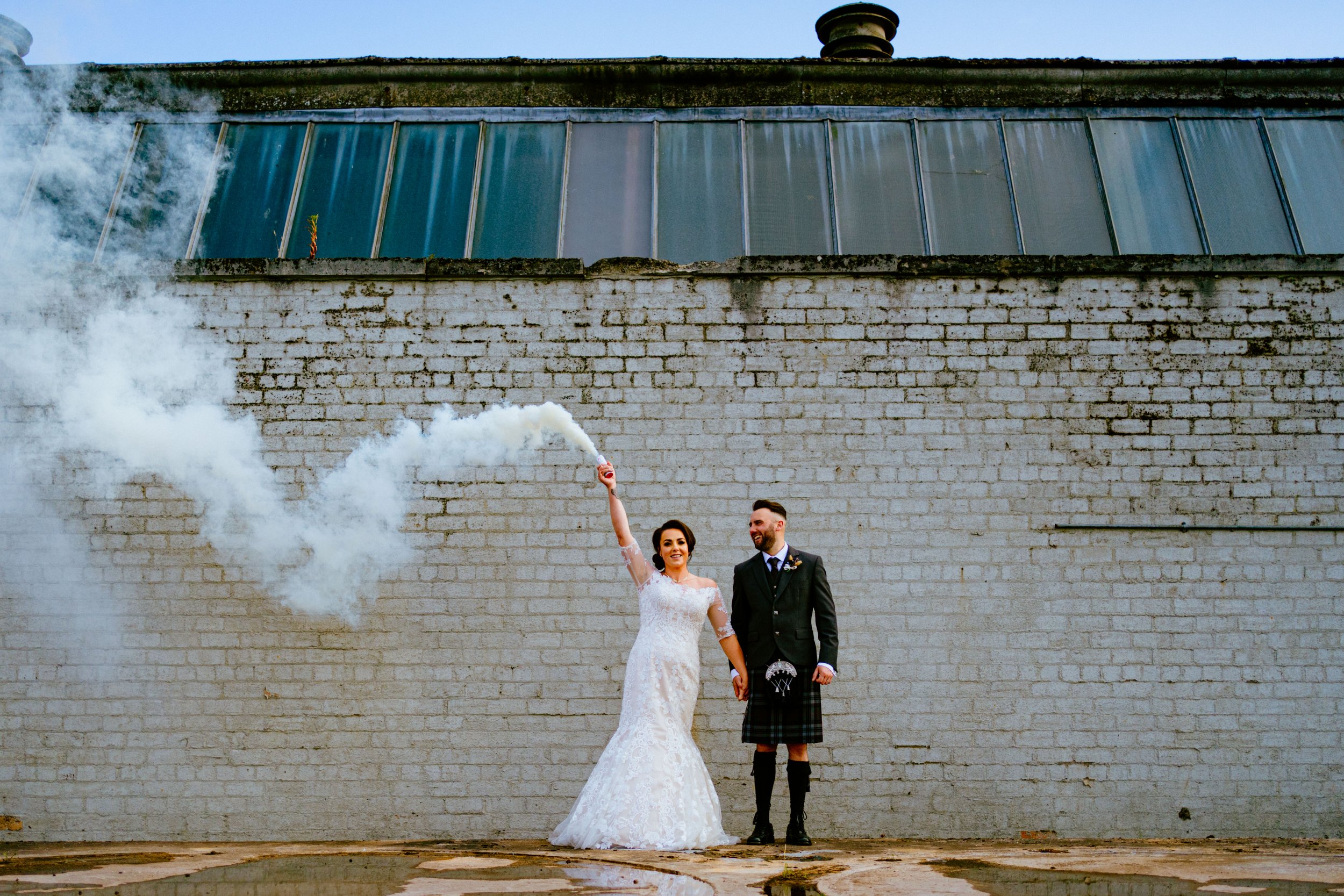 Wedding couple at The Weaving Shed in Dundee, standing against an industrial brick wall as the bride holds a smoke flare, creating a bold and playful moment.