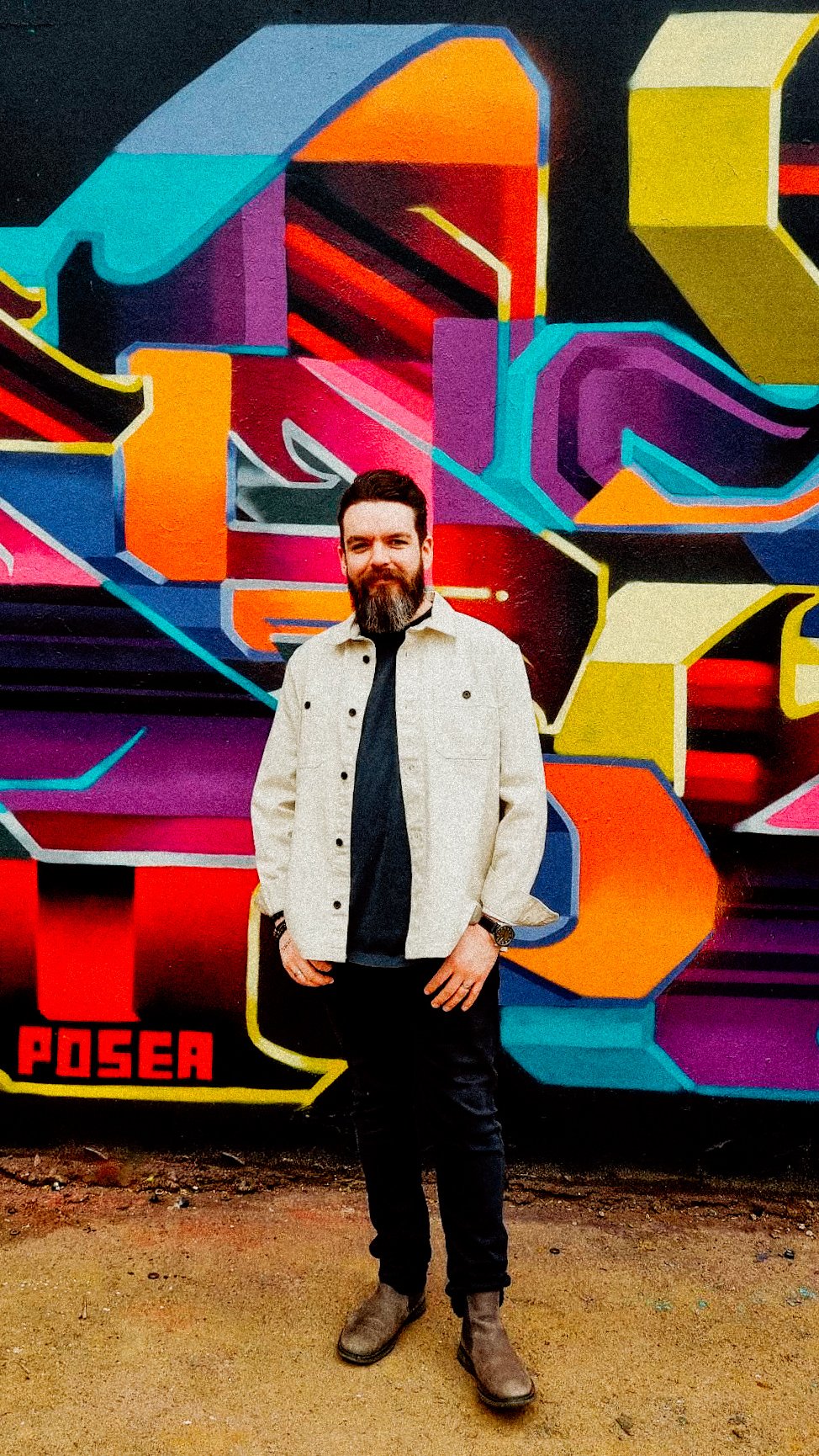 Portrait of a bearded man standing at Yardworks Festival in Glasgow, in front of colourful graffiti artwork by Posea.