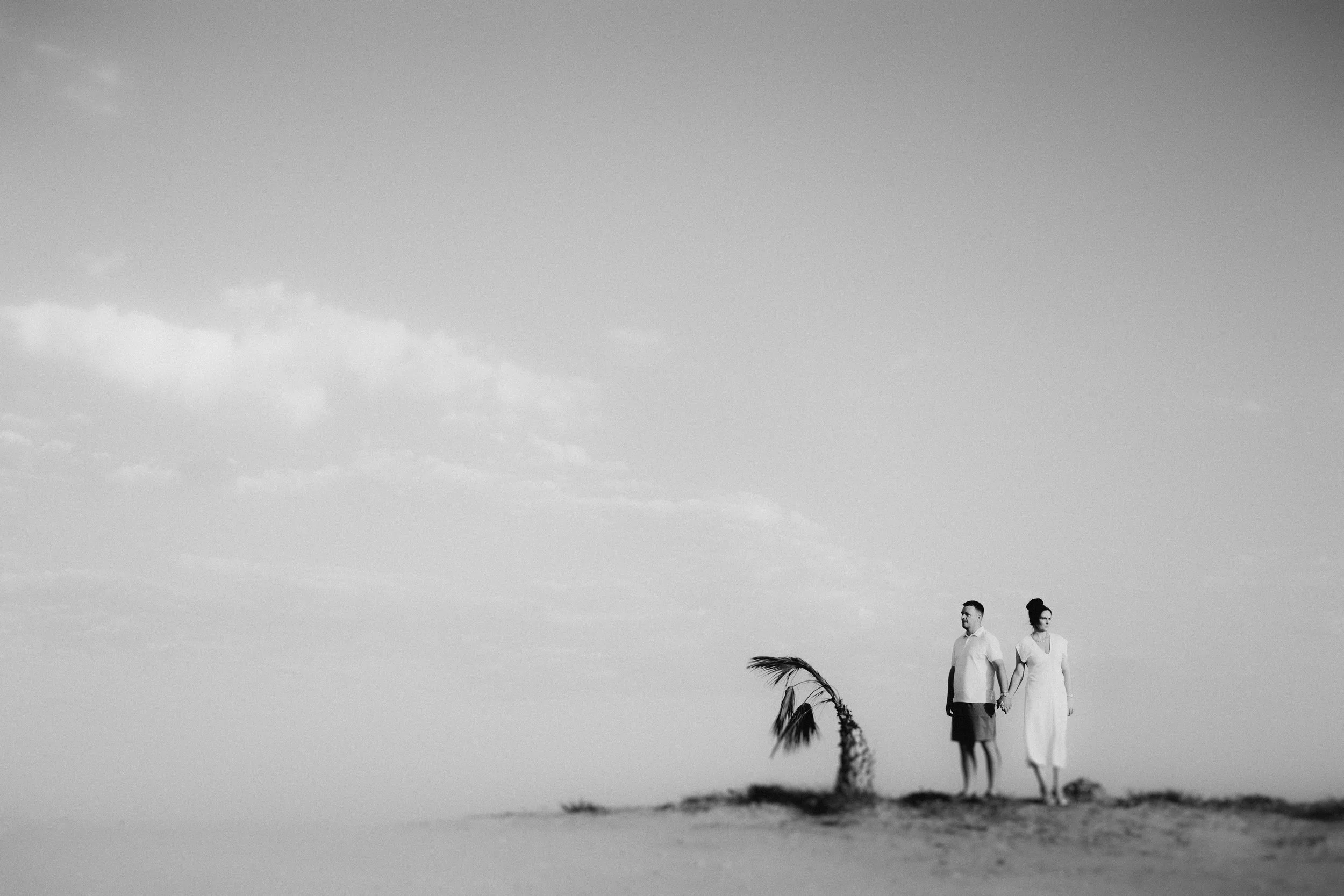Pre-wedding shoot in Paphos, Cyprus, with a couple holding hands on a beach beside a windswept palm tree, captured in a minimal black and white composition.