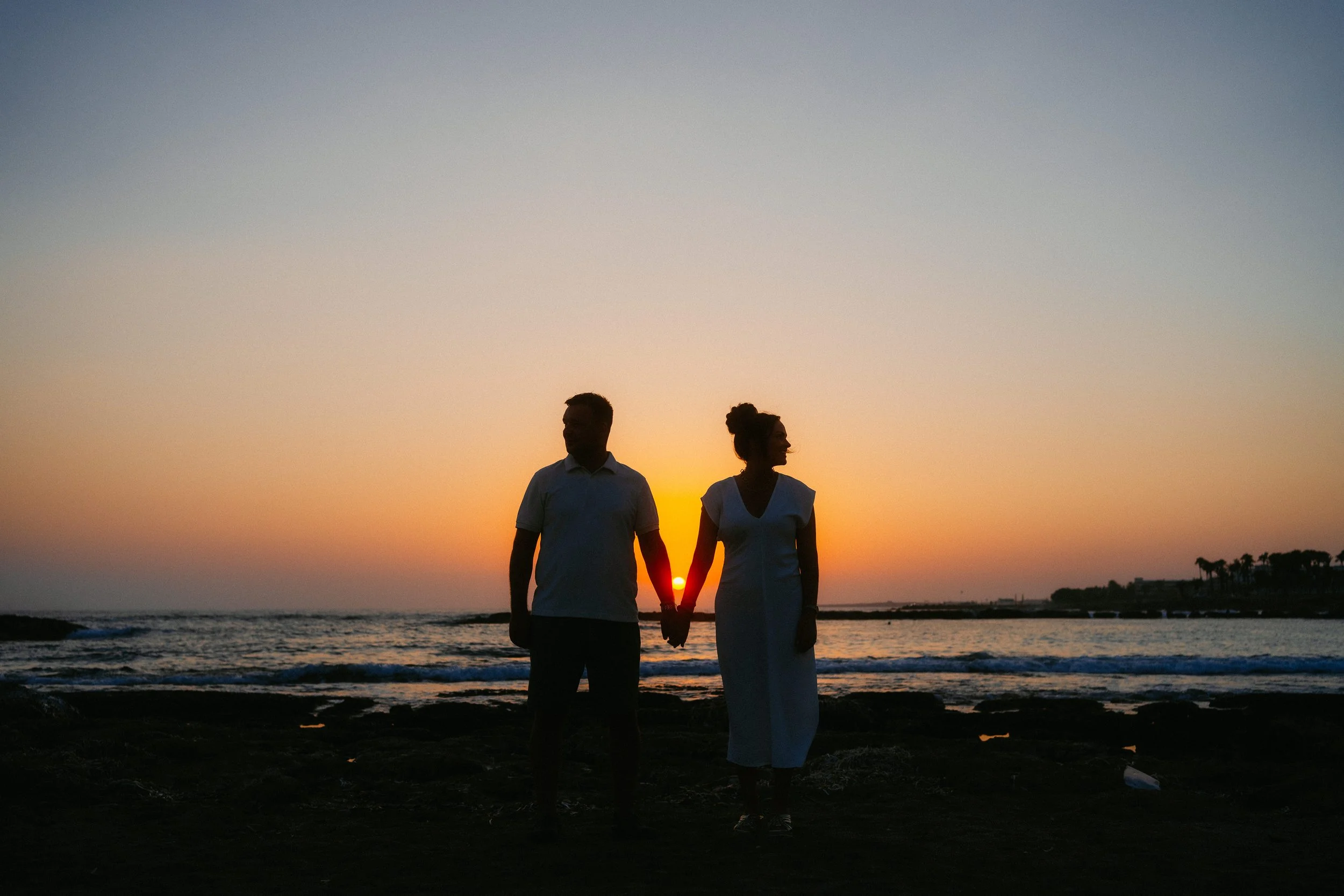 Pre-wedding shoot in Paphos, Cyprus, with a couple holding hands on the shoreline at sunset, silhouetted against the sea and warm evening sky.