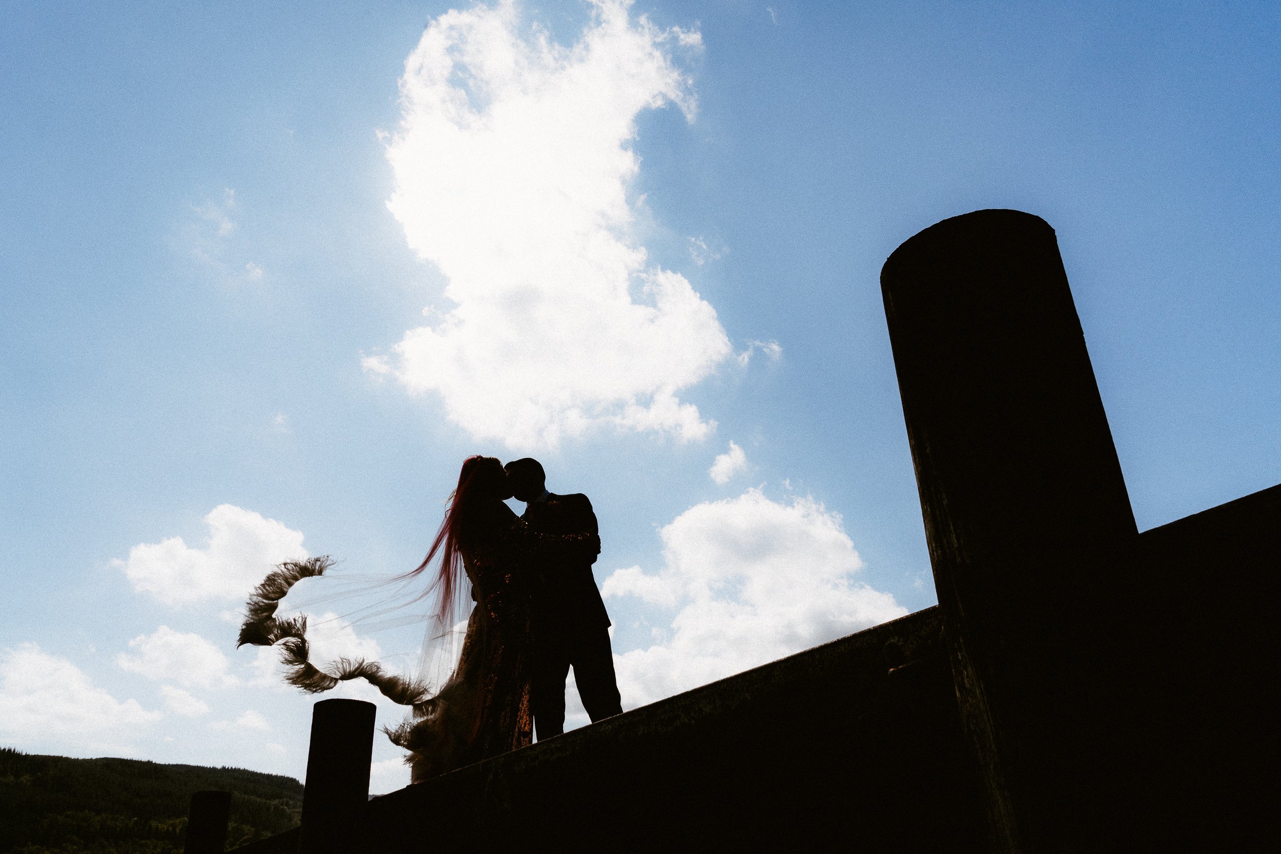 Loch Venachar wedding couple silhouetted on a pier, kissing against a bright sky with flowing veil and dramatic landscape in the background.