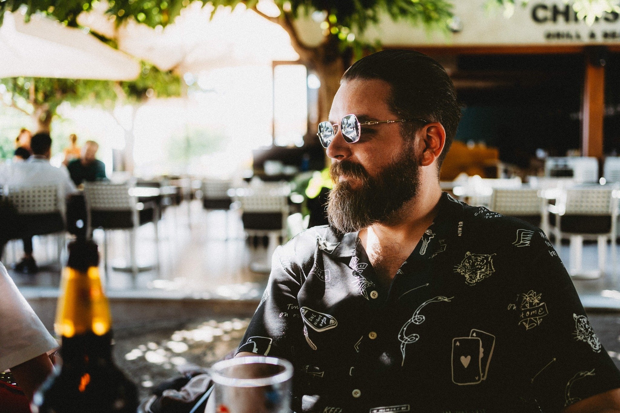 Portrait of a bearded man wearing sunglasses, sitting outdoors in Paphos, Cyprus, enjoying a relaxed moment with a drink in warm natural light.