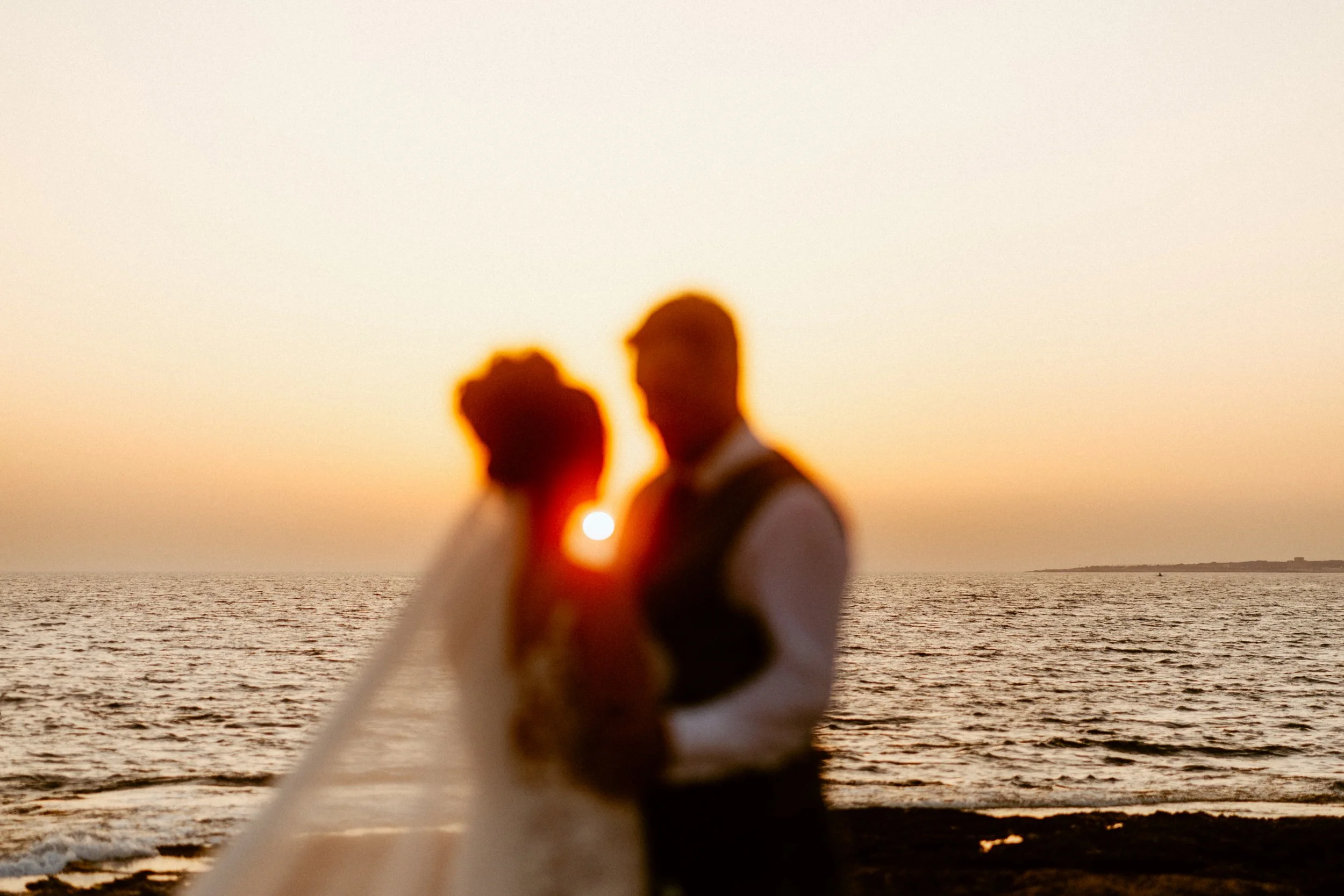 Wedding in Paphos, Cyprus, with a couple softly out of focus as they stand together by the sea at sunset, the sun setting between them.