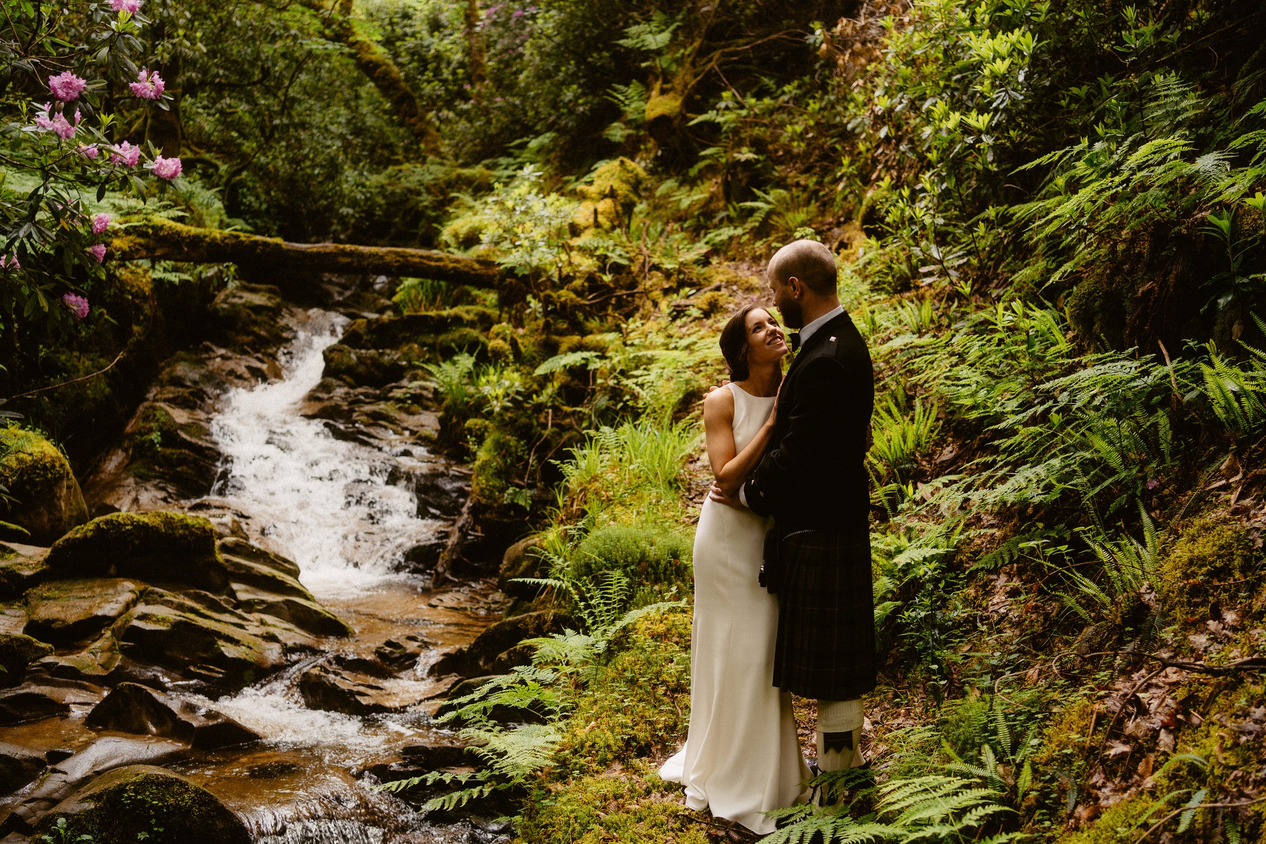 Wedding couple in the gardens of Stuckgowan House, standing beside a woodland stream and waterfall, sharing a relaxed moment after their ceremony at Fruin Farm.