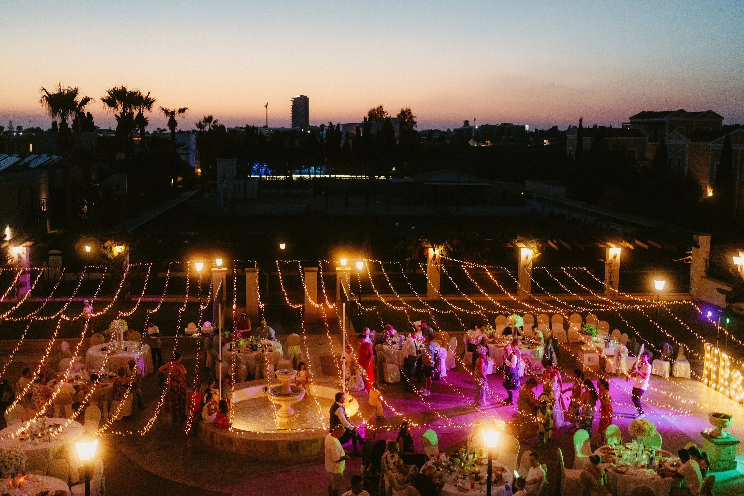 Paphos wedding in Cyprus with an outdoor evening reception, featuring guests dining and dancing under string lights at sunset.