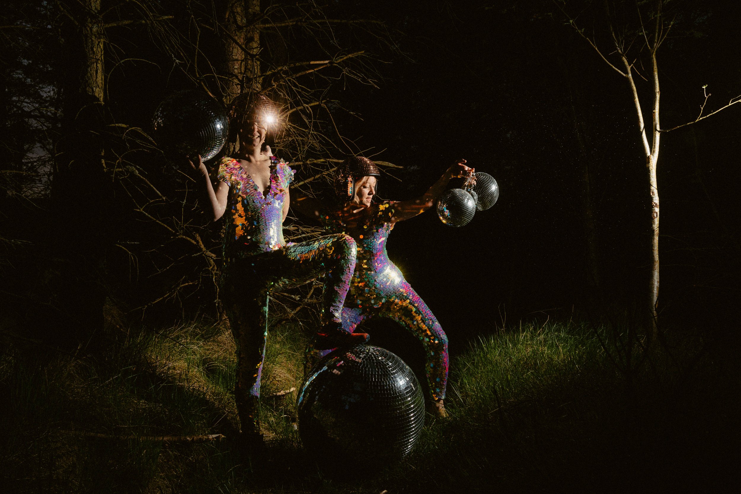 Engagement shoot in Glencoe at night. Couple wearing colourful sequin outfits pose in a forest holding disco balls, lit dramatically against a dark woodland backdrop.