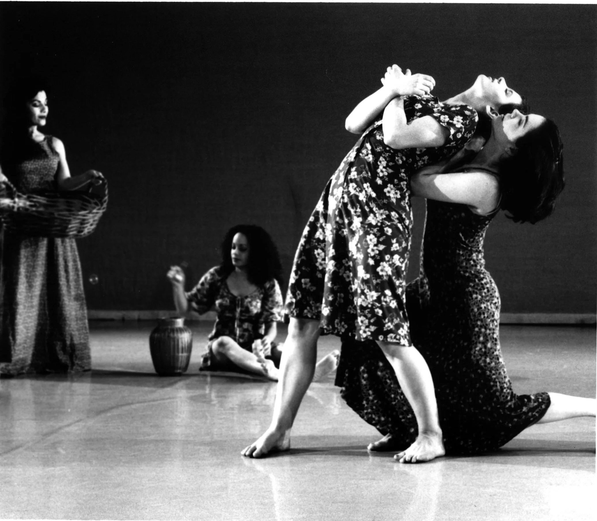 Black and white photo of two women practicing yoga on a stage, with one woman upside down on the other in a partnered pose, while another woman sits with her eyes closed and hands in a prayer position in the background.