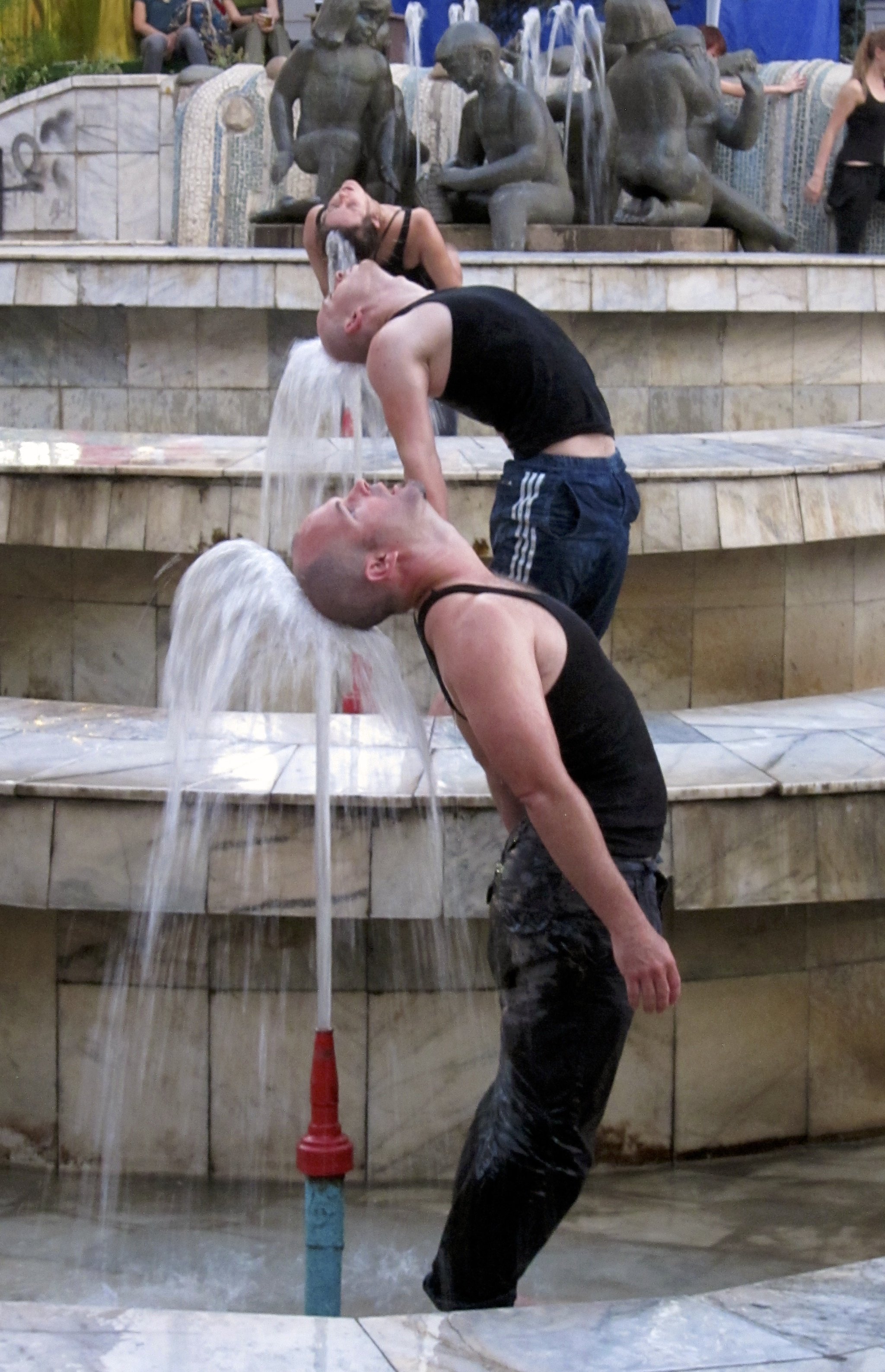 Three bald men in tank tops leaning back under flowing water from fountains at a public space, with a woman in the background.