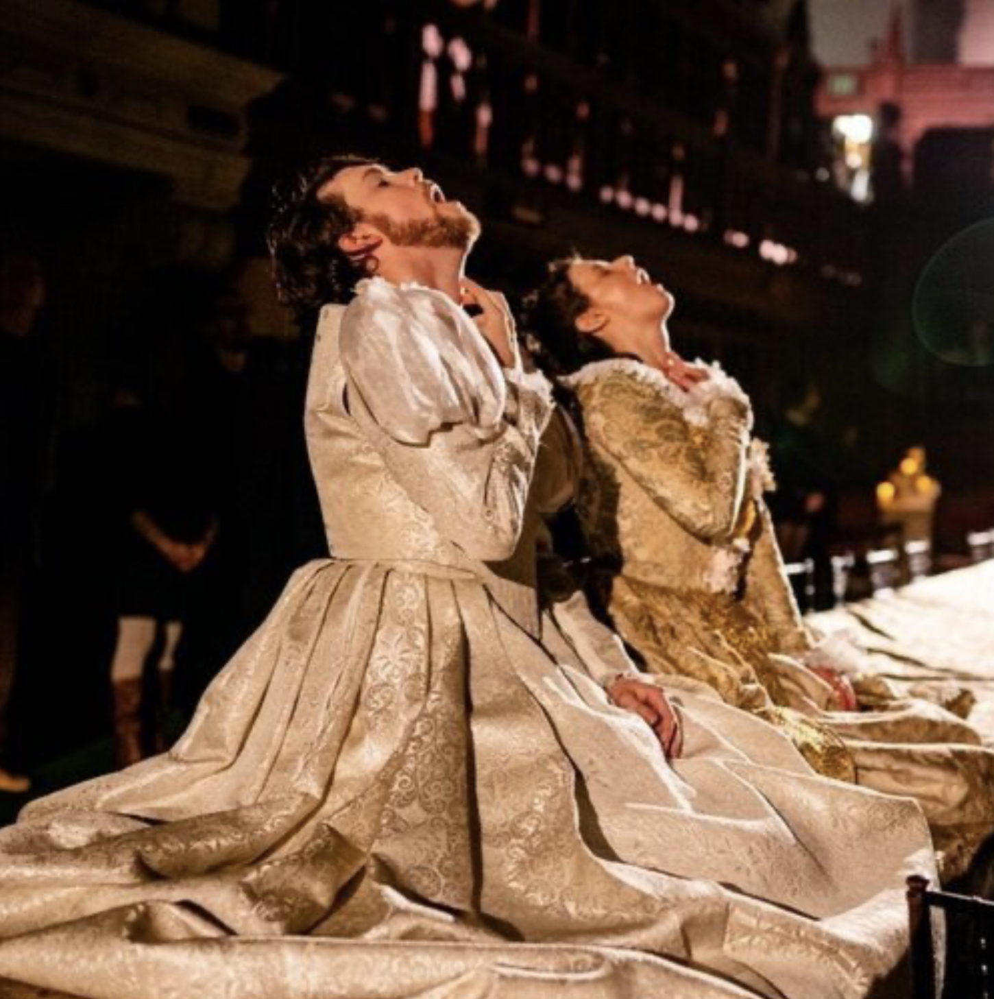 A man and a woman in ornate, gold-embossed dresses kneeling on a banquet table with open mouths, with their heads tilted upwards and eyes closed, in a dimly lit library reading room, with a warm glow.