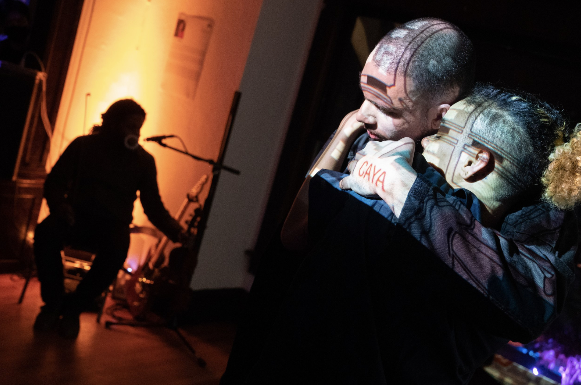 Two people embracing at a dimly lit event or performance, with a musician in the background playing guitar, and orange ambient lighting.