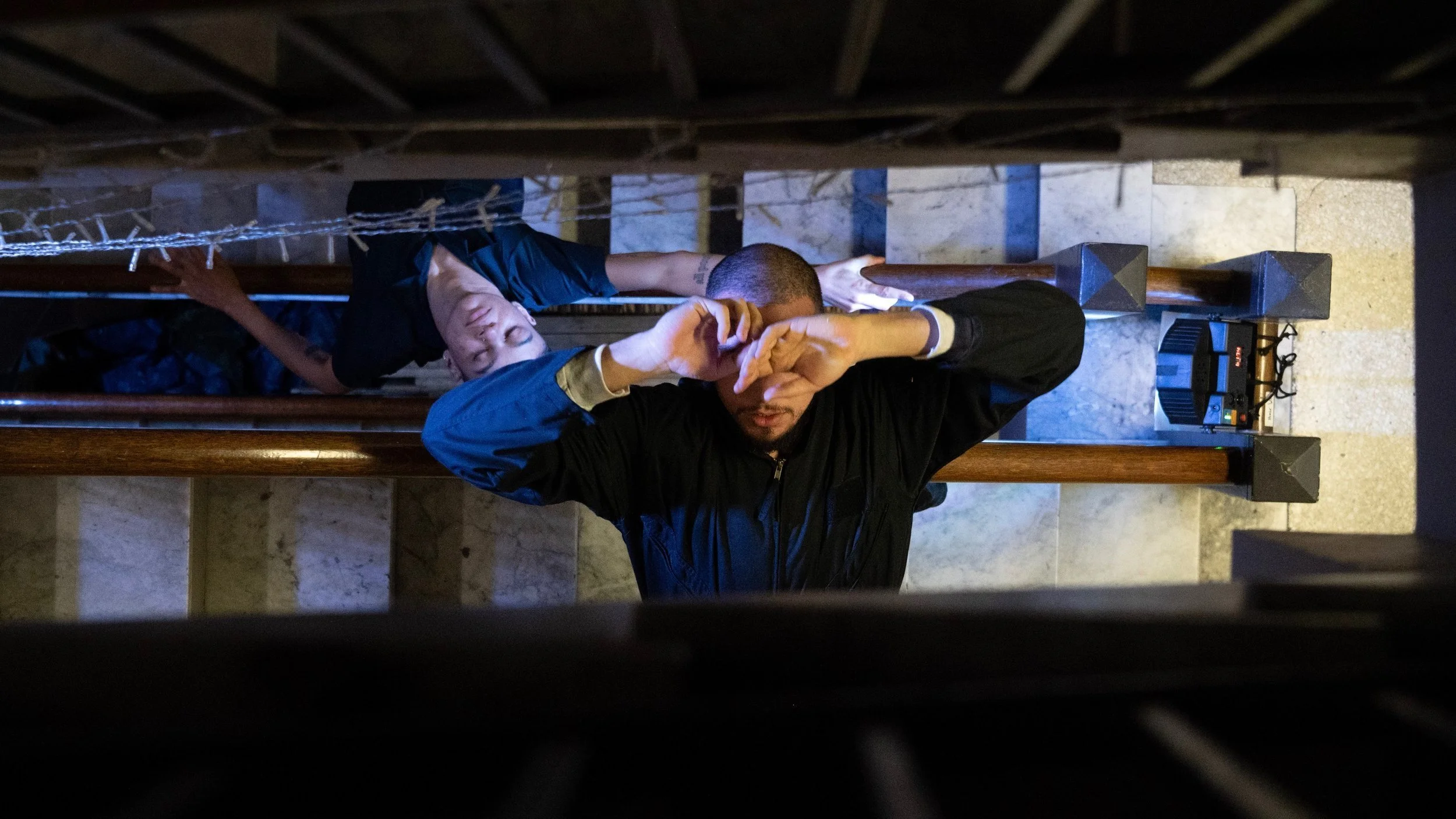 Two dancers laying across stair banisters, one with his hands covering his face and the other lying with eyes closed, seen through a staircase railing.