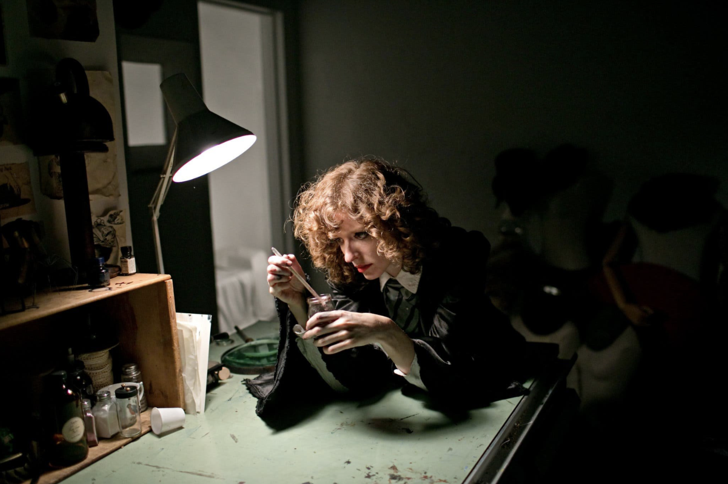 A woman with curly red hair sitting at a cluttered worktable in a dimly lit room, working on an art project using a glass jar and a paintbrush, with a lamp illuminating her workspace.
