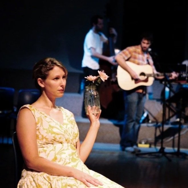 A pregnant woman with blondish brown hair in a yellow patterned dress sitting on a chair, holding a glass jar with pink flowers, in front of a stage with two men performing music, one singing with a guitar and the other playing a bass guitar.