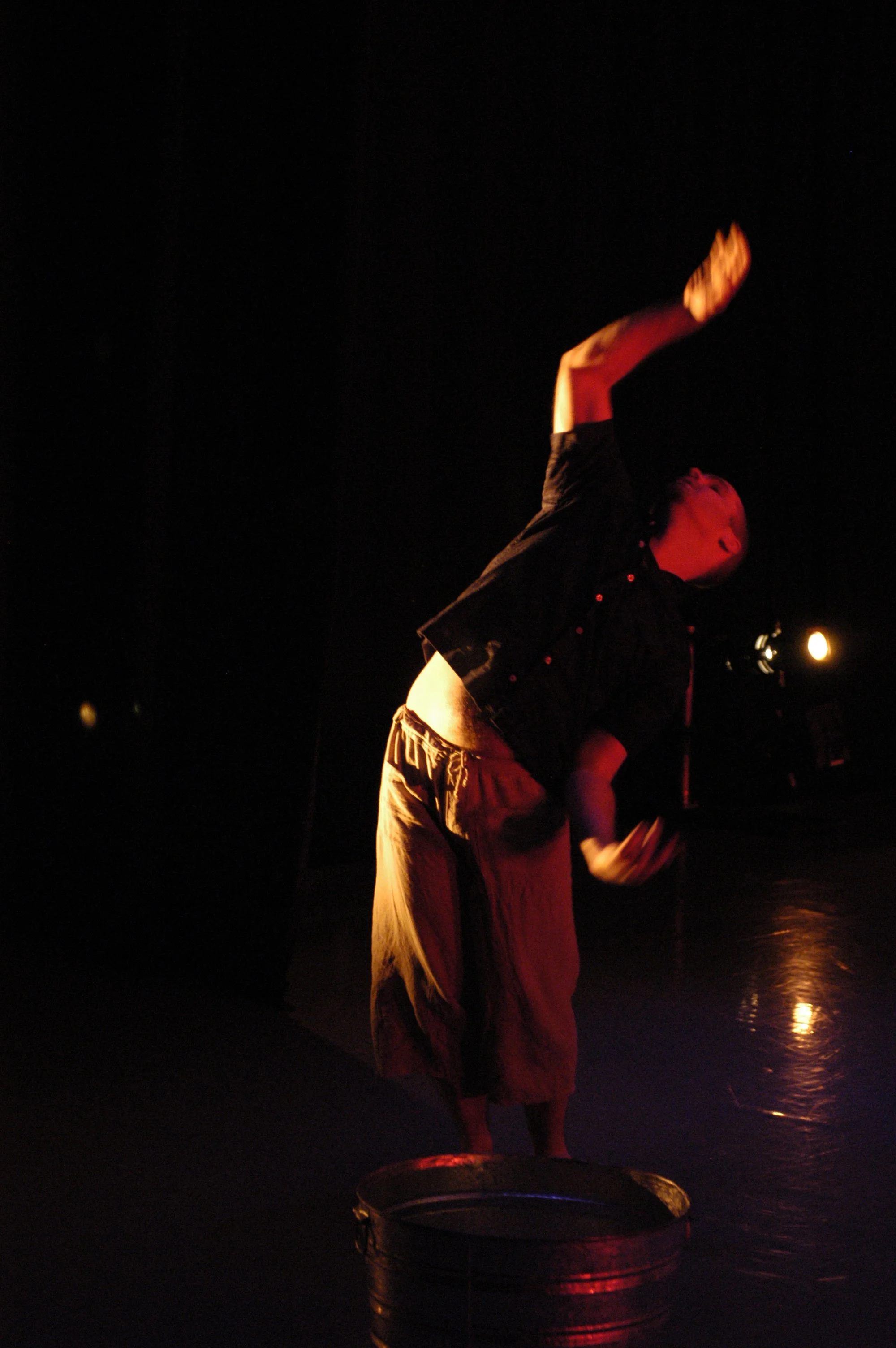 Person on stage at night, wearing loose pants and a dark shirt, with one arm raised, holding a book or paper, illuminated by warm lighting, with a dark background and reflections on a wet surface.