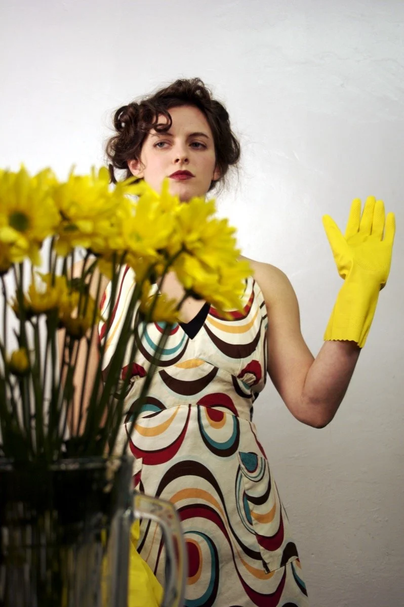 A woman holding a bouquet of yellow flowers looking to her left, with a person behind her raising their legs and feet near her head against a white wall.