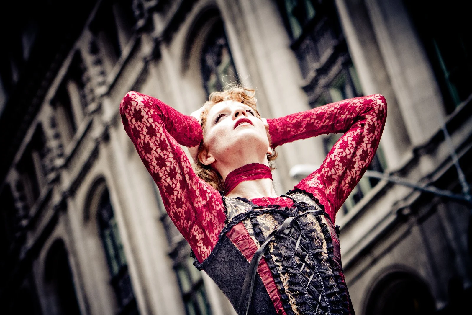 A woman with short red hair wearing red lace clothing and a choker, looking upward with her arms raised behind her head, against a blurred urban street background.