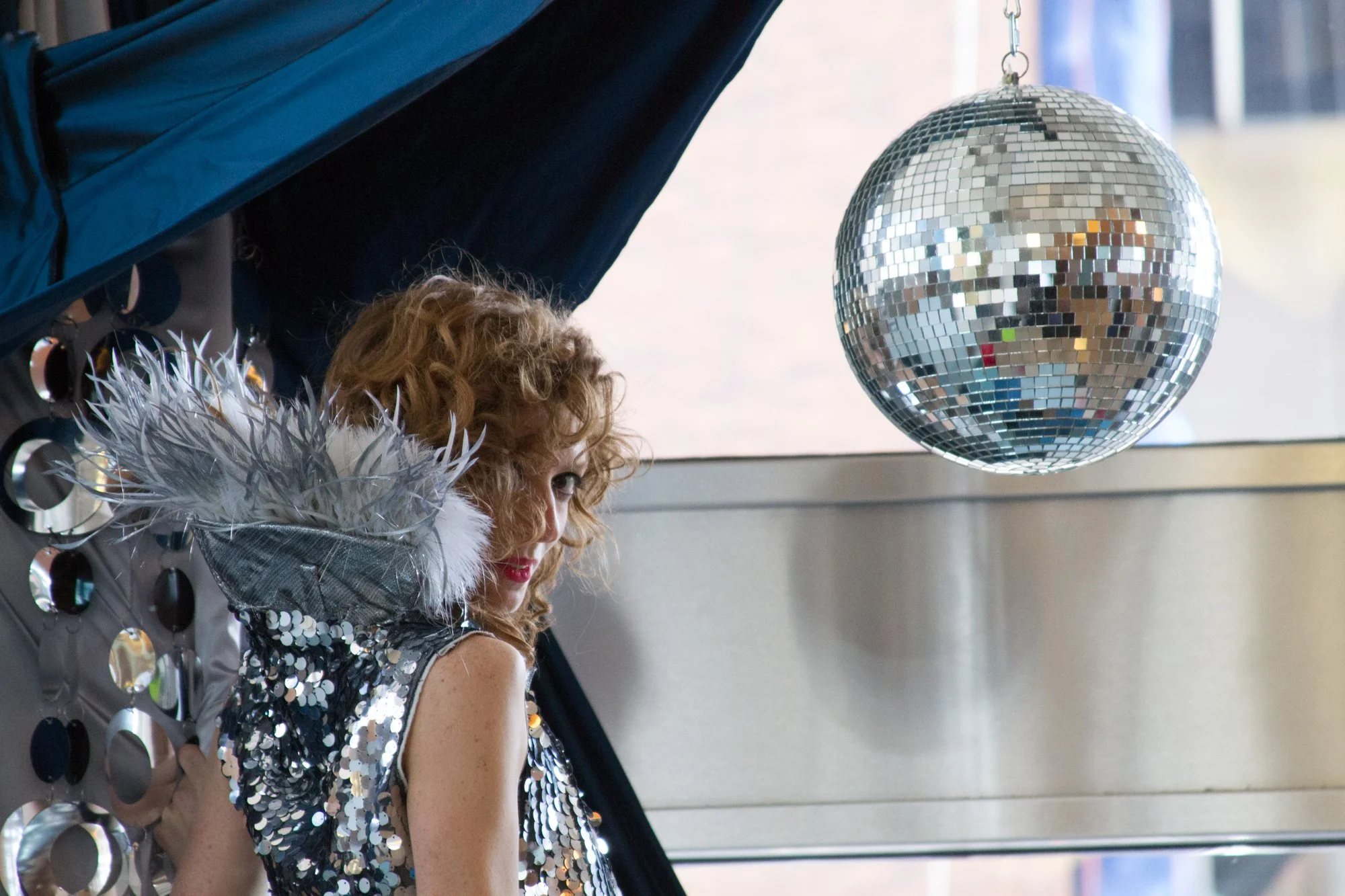 A woman with curly hair wearing a silver sequin dress and a feathered shoulder accessory, standing near a metallic wall with circular decorations.