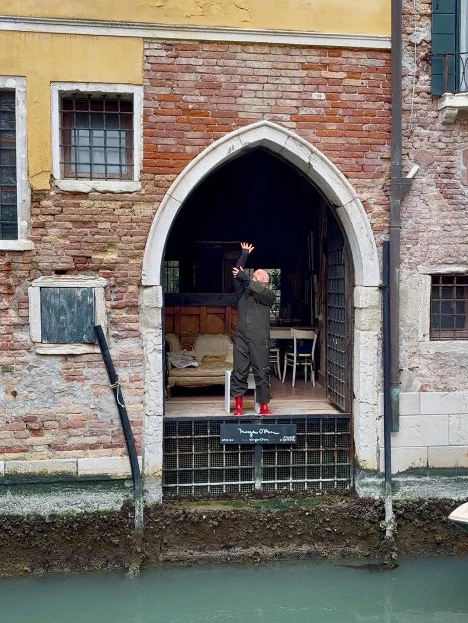 A person with a bald head standing on a platform in front of a building with brick walls and arched windows, miming a gesture as if holding an invisible object, with water below and a partly submerged drainpipe nearby.