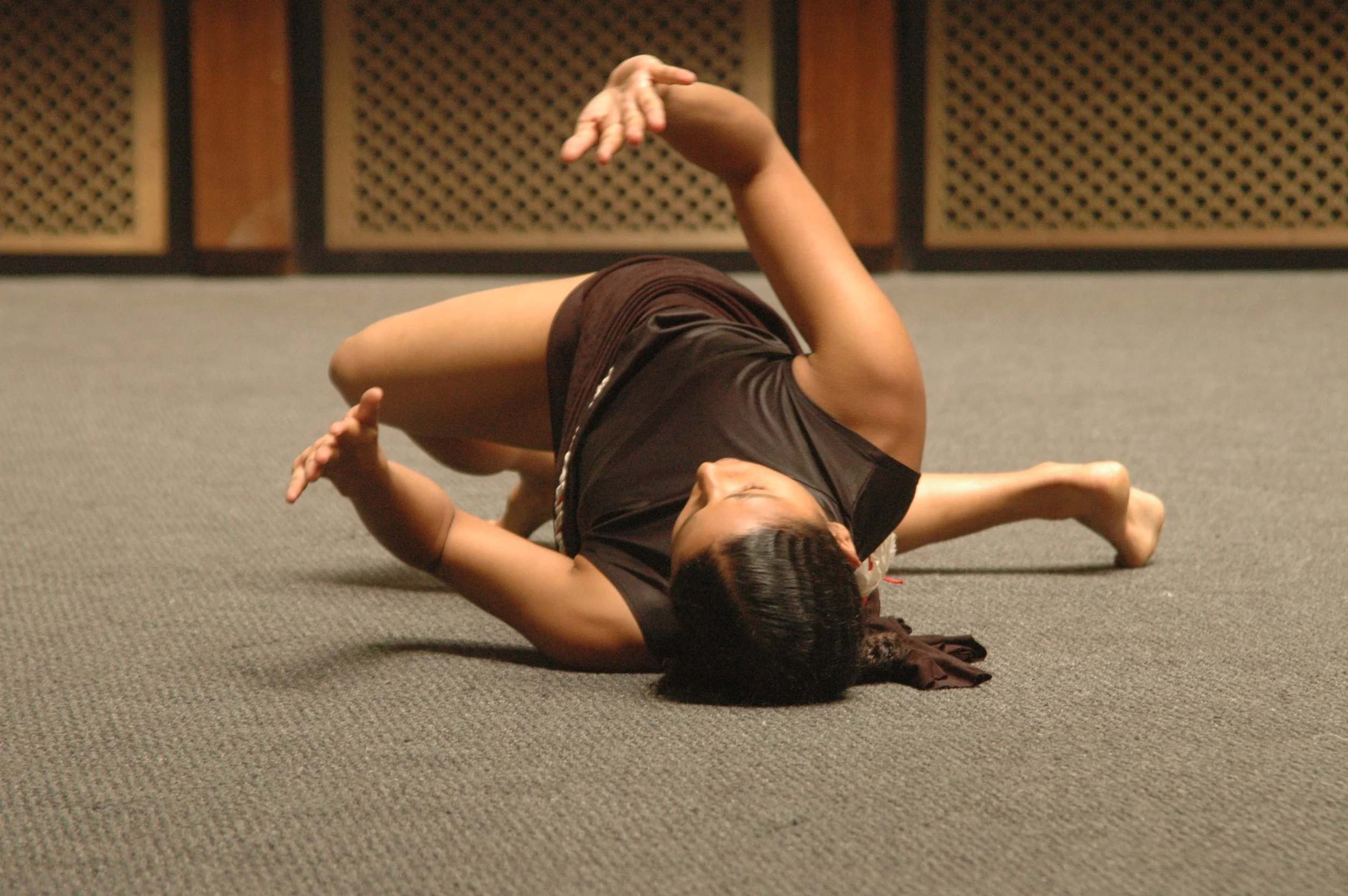 A person lying on the floor in a pose that resembles a frog, with their elbows bent and hands extended in front, in a room with a gray carpet and wooden panels with a lattice pattern in the background.