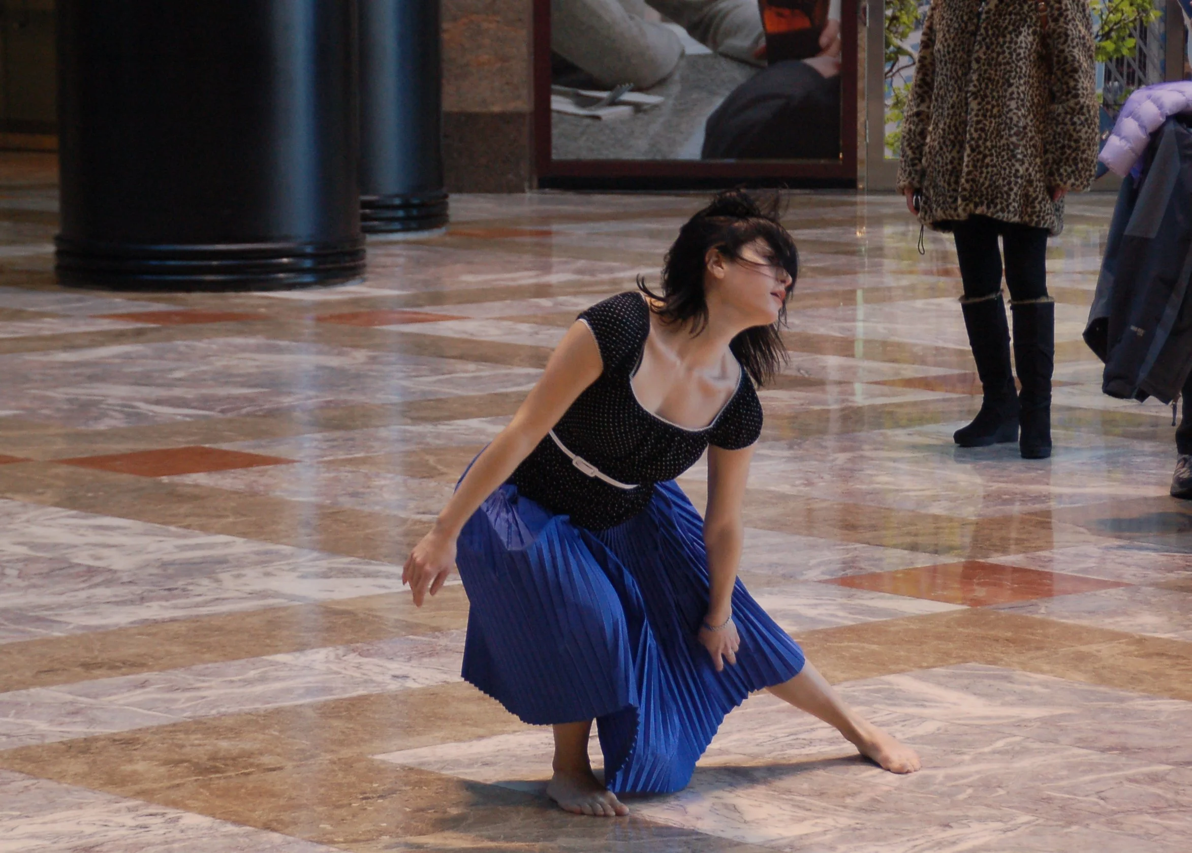 A woman with black hair, glasses, and a black top with white dots is performing a dance move on a marble floor. She wears a long blue pleated skirt and is barefoot. In the background, there are two people sitting at a table, one holding a smartphone, and another standing nearby wearing a leopard print jacket and black pants. The setting appears to be indoors, possibly in a lobby or auditorium.
