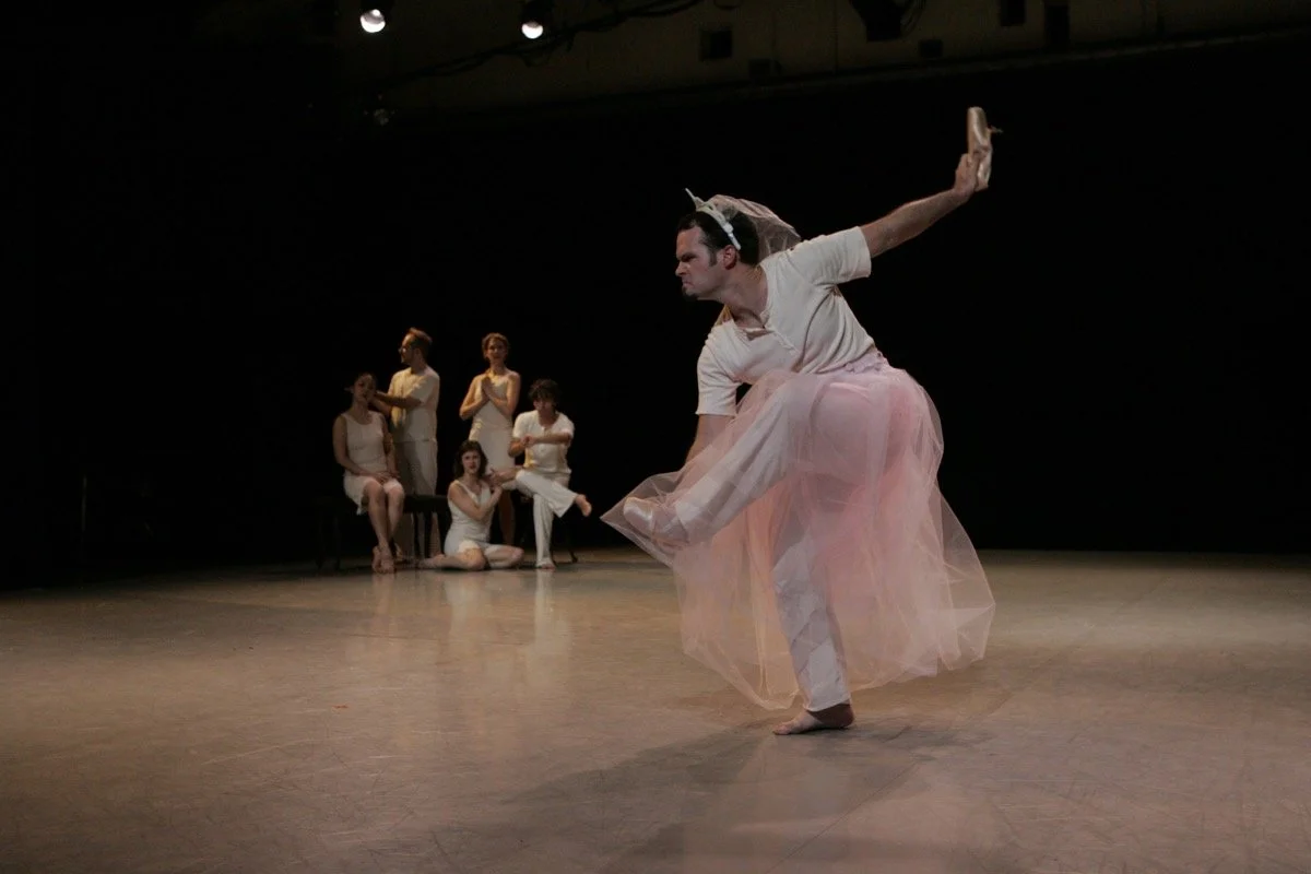 A ballet dancer in a pink tutu and white shirt performing on stage, with four women watching in the background.