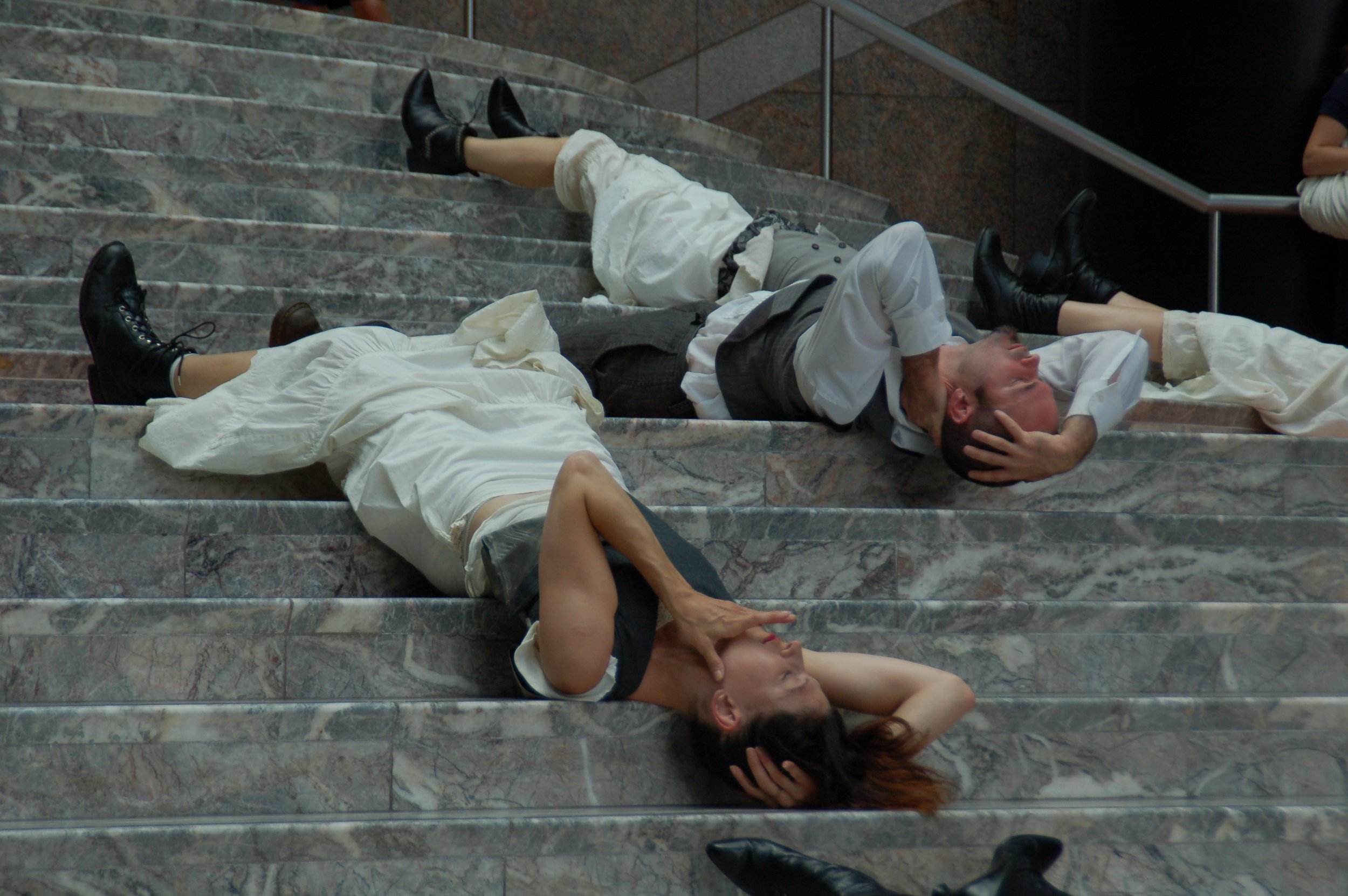 Three people lying on marble stairs, appearing to be unconscious or resting, with their hands on their heads and bodies relaxed.
