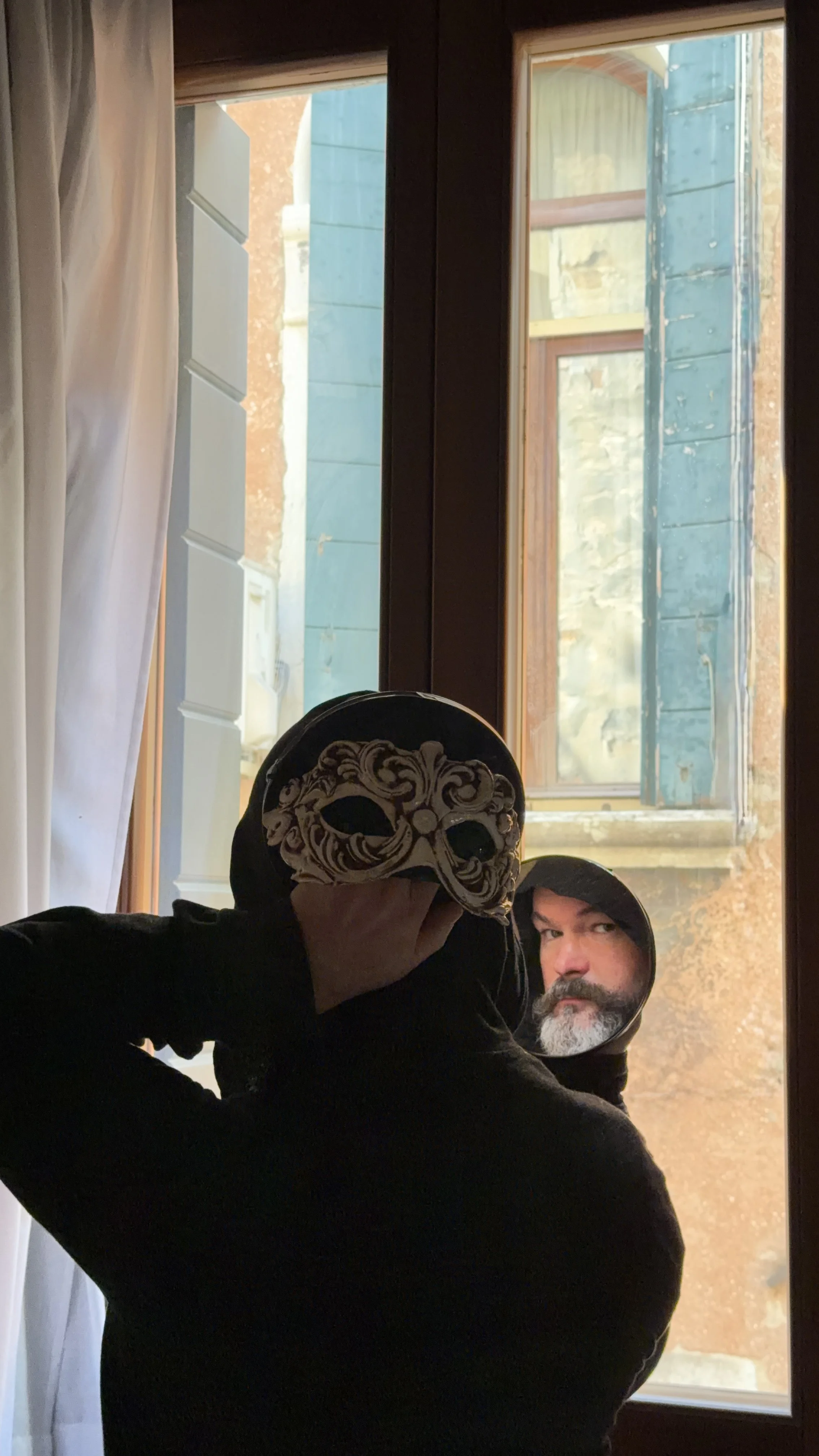 A person taking a mirror selfie, holding a Venetian mask in front of their face, with a window and outdoor scene reflected in the mirror. The person has a beard and is wearing a hoodie.