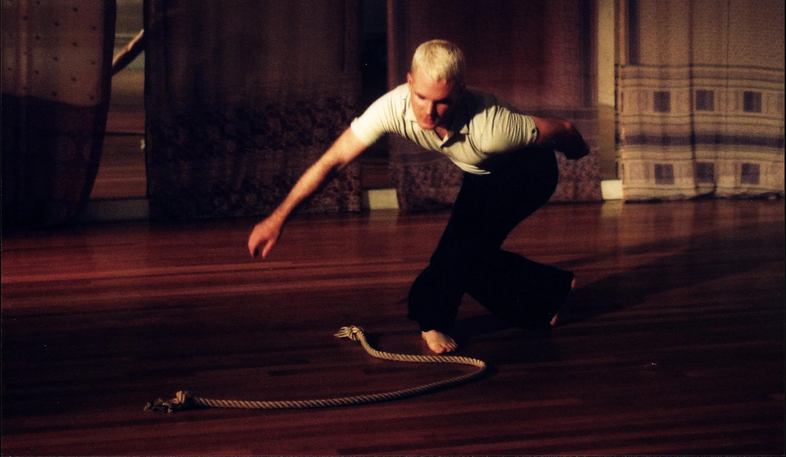 A person with blonde hair crouching on a polished wooden floor, reaching towards a rope lying on the floor, with dark wooden paneling in the background.