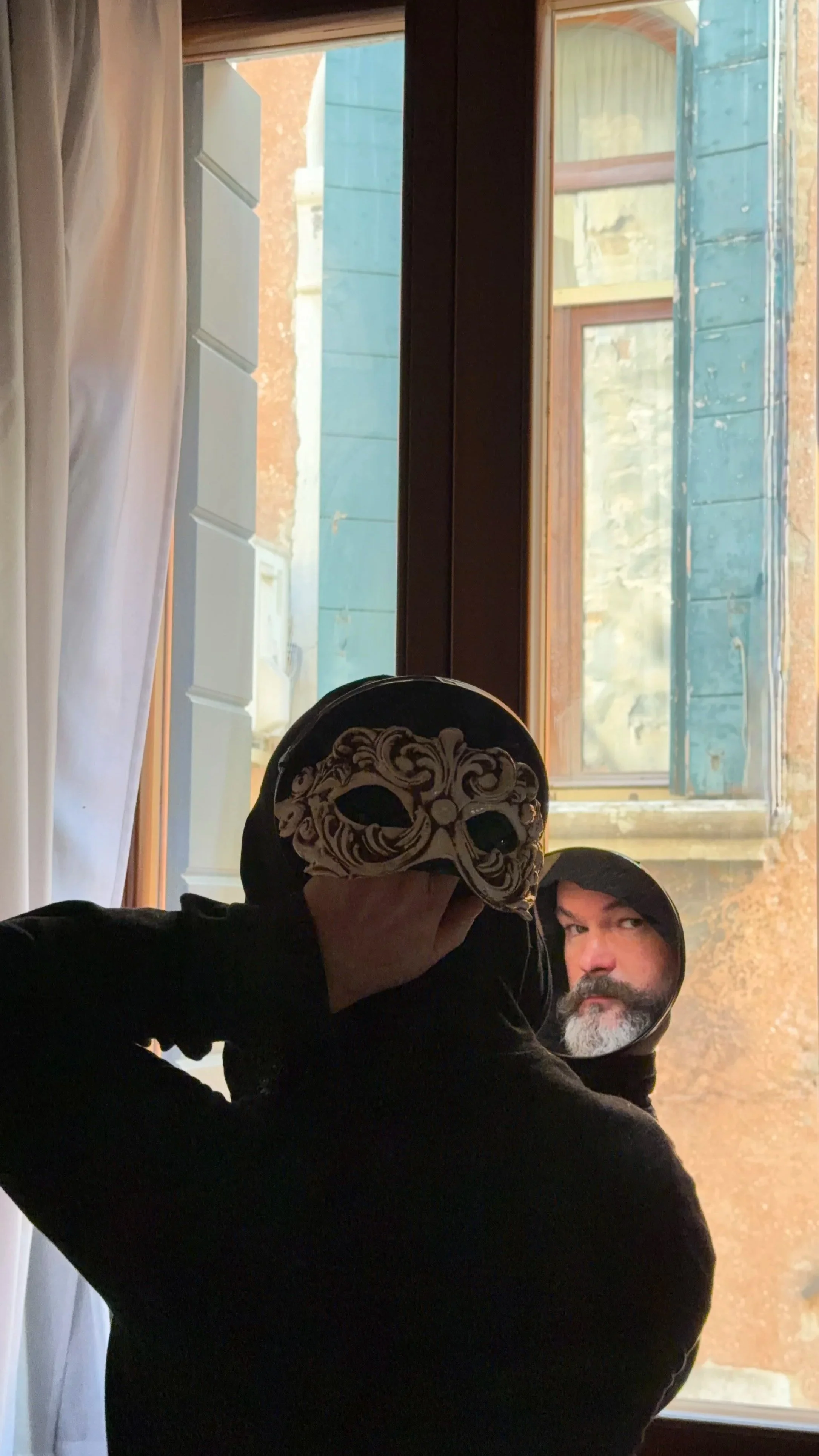 A person taking a mirror selfie, holding a Venetian mask in front of their face, with a window and outdoor scene reflected in the mirror. The person has a beard and is wearing a hoodie.
