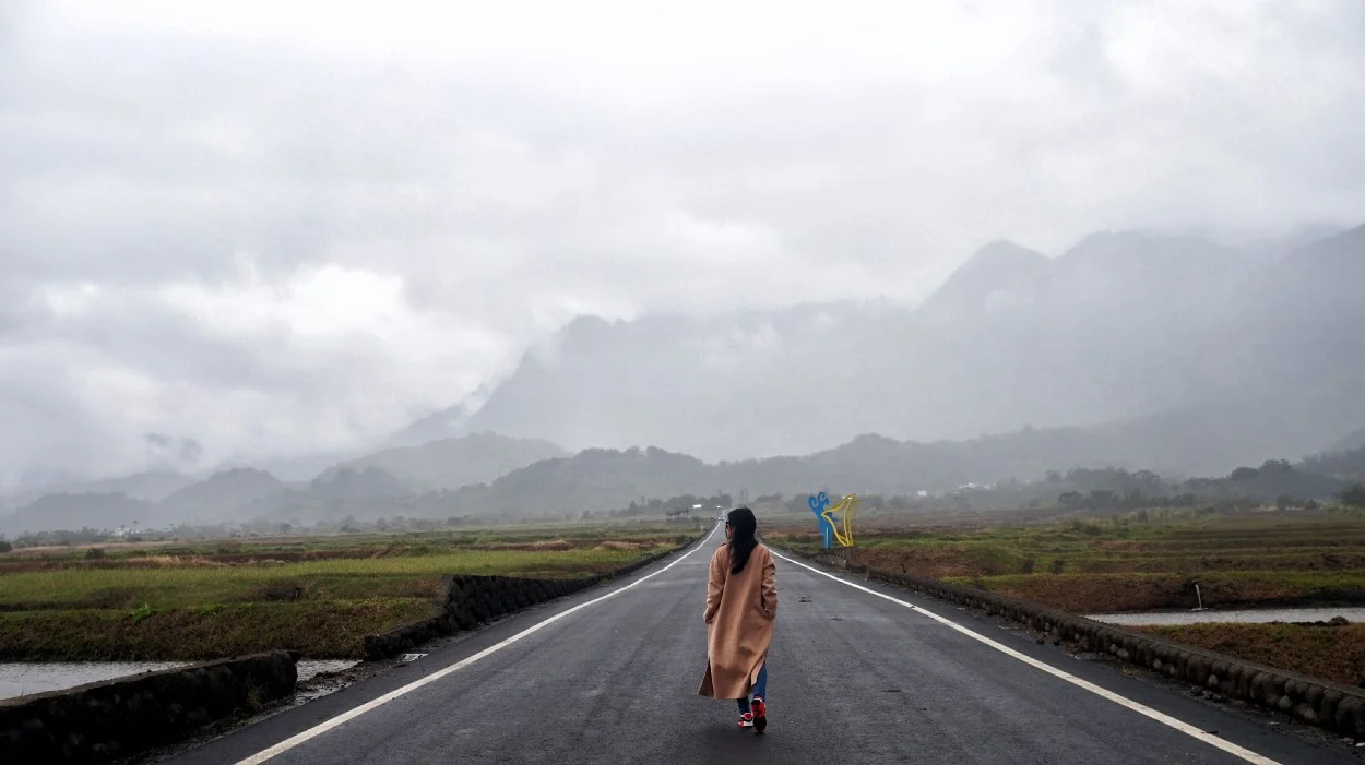 woman with long coat walking down middle of road towards mountains