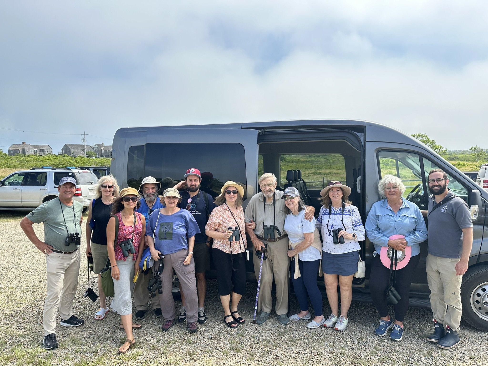 picture of a group of 12 people on a birding trip standing in front of a van, they have binoculars