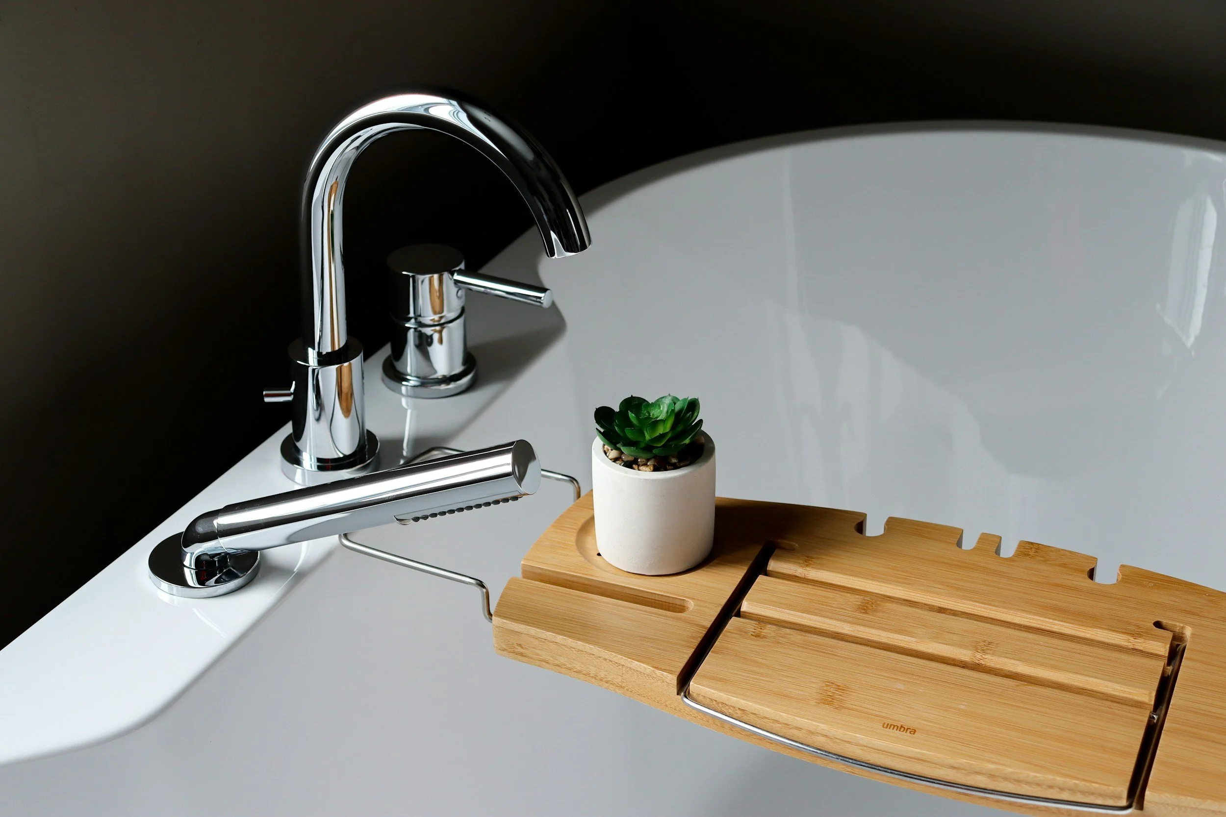 A white bathtub with a modern chrome faucet, a small potted succulent plant, and a wooden bath tray.