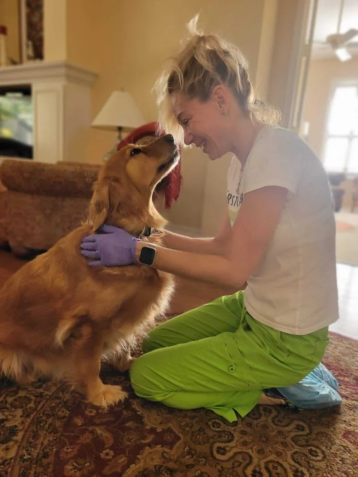 Woman smiling and kneeling on the carpet, touching foreheads with a golden retriever dog, inside a cozy living room.