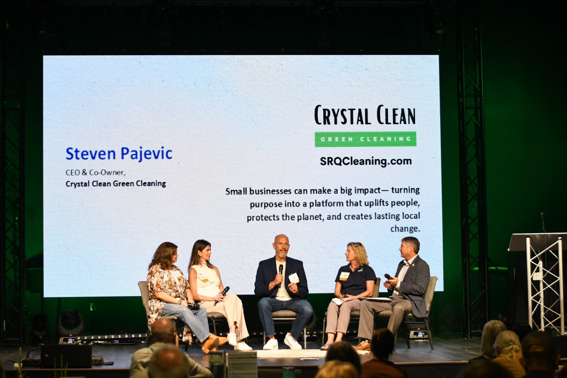 Panel discussion at a conference with five individuals seated on stage, large screen behind them displaying a presentation slide with the title 'Crystal Clean Green Cleaning' and the name Steven Pajevic, CEO of Crystal Clean Green Cleaning.