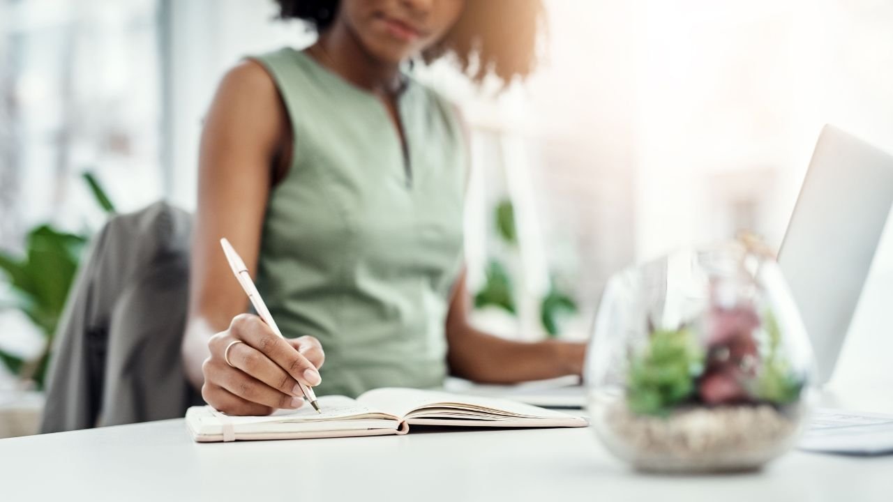 A woman in a green sleeveless top writes in a notebook at a desk, with a glass bowl of succulents and a laptop nearby.