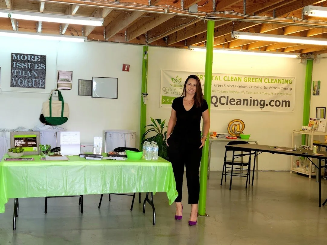 A woman standing inside a green-themed office or event space with tables, chairs, and signage promoting green cleaning services.