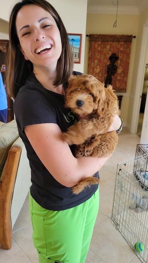 A woman with brown hair holding a small, fluffy, brown puppy and smiling in a home interior.